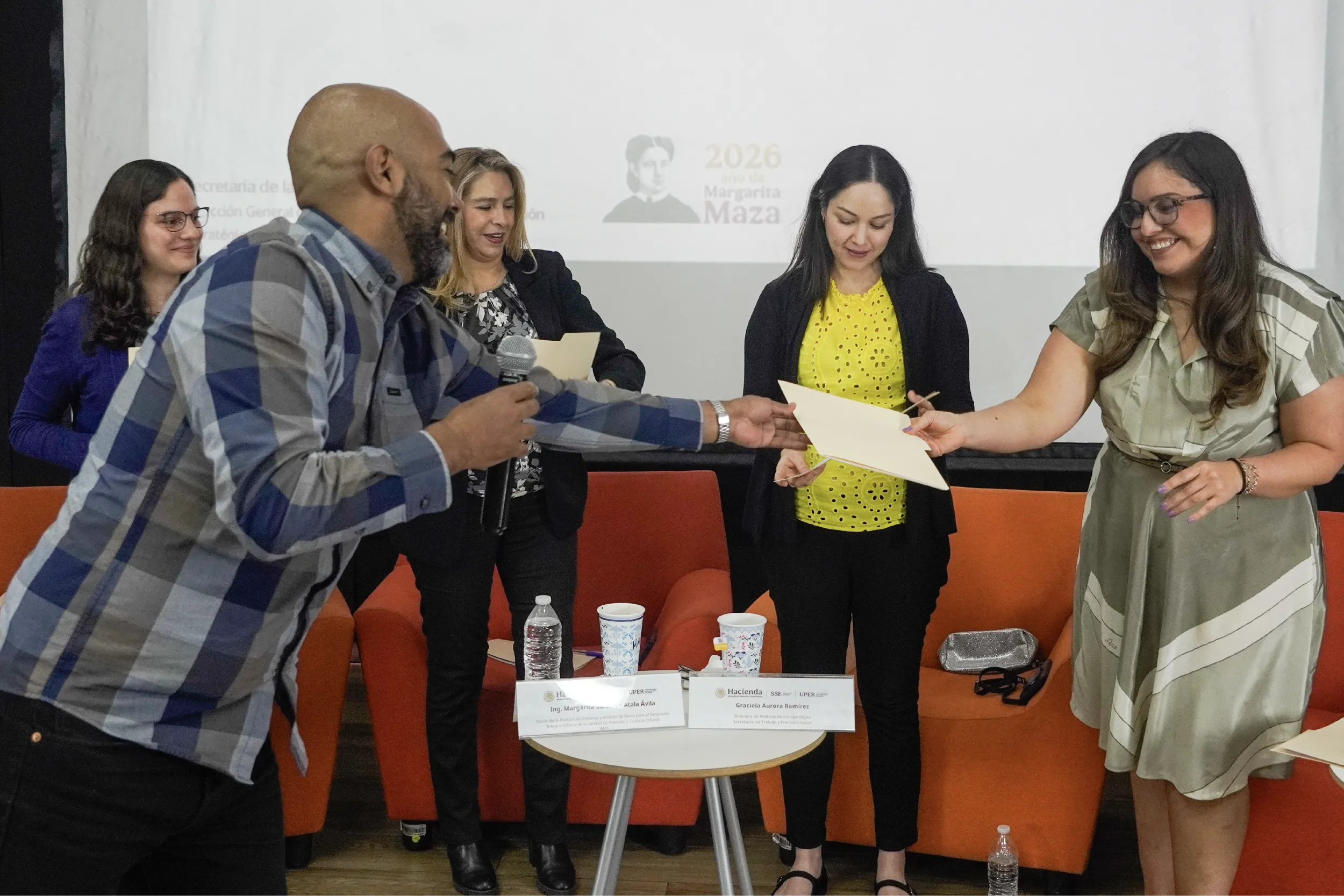 Man in plaid shirt hands a certificate to a woman in a bright yellow top as others smile and observe, in a panel setting with name tags on a table and water cups nearby.