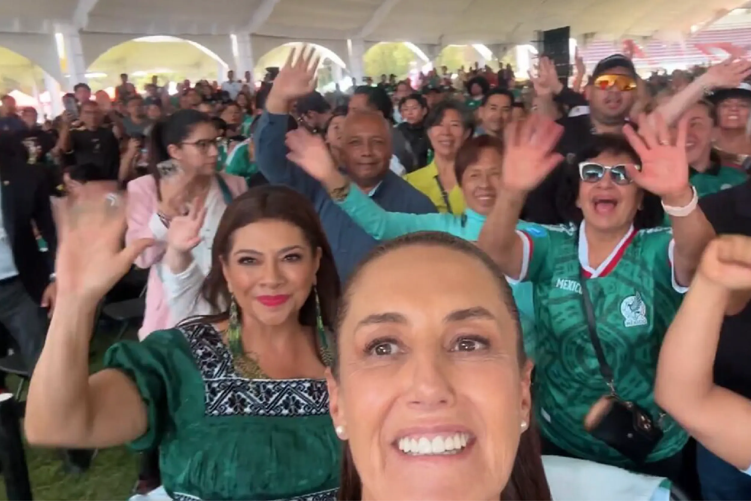 Crowd selfie at an outdoor event under a tent, group waving at the camera with a smiling woman in green in the foreground