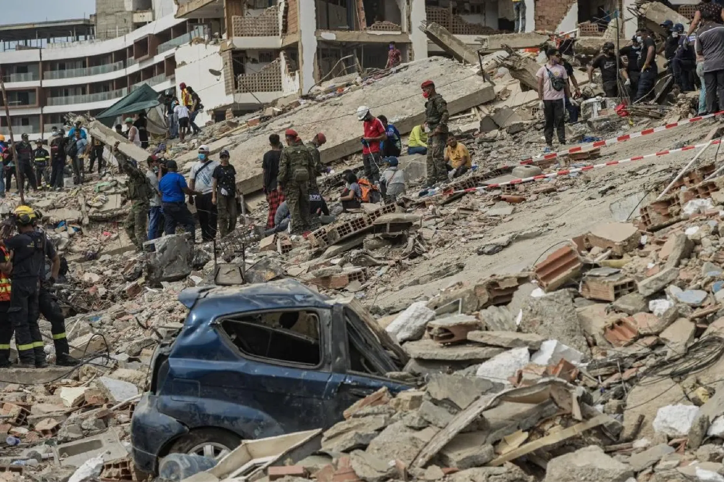 Rubble-strewn street with rescue workers and civilians amid collapsed buildings and a crushed blue car in the foreground.