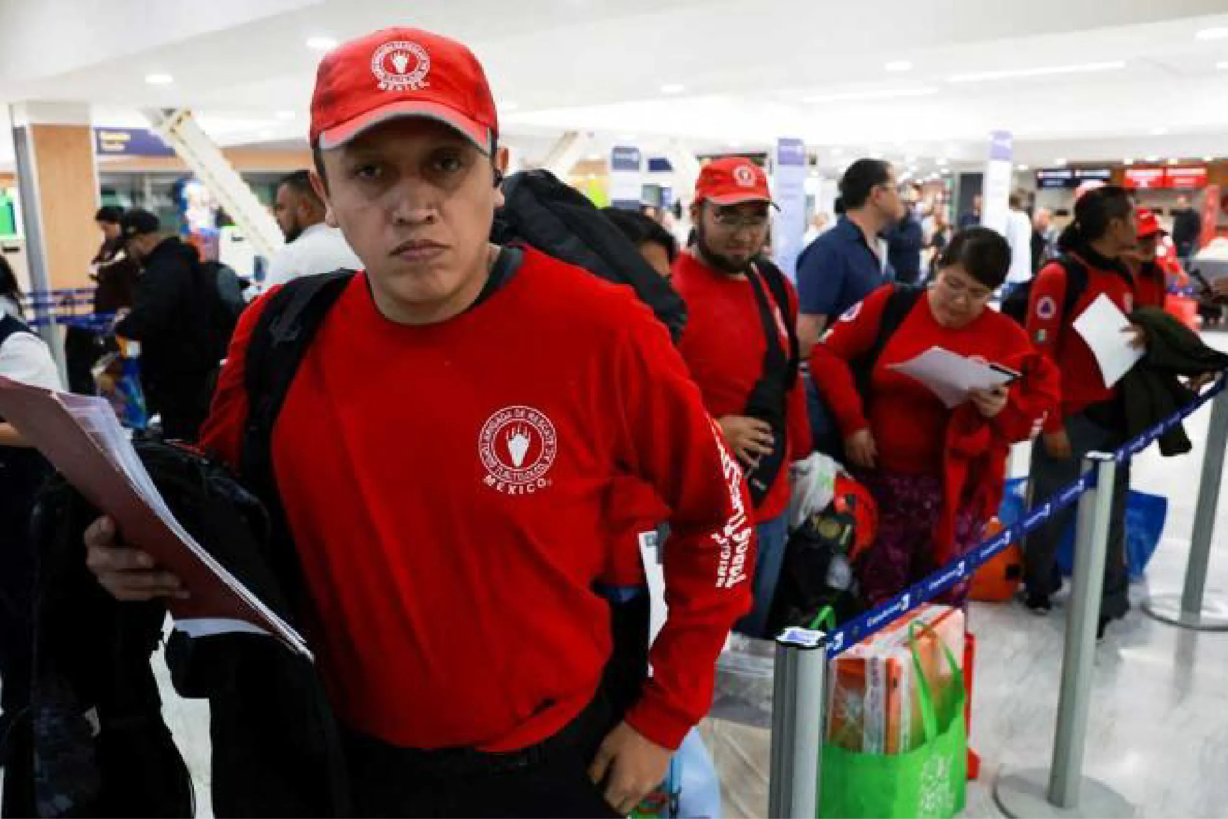 Group of volunteers in red shirts and caps queuing at an airport check-in area.