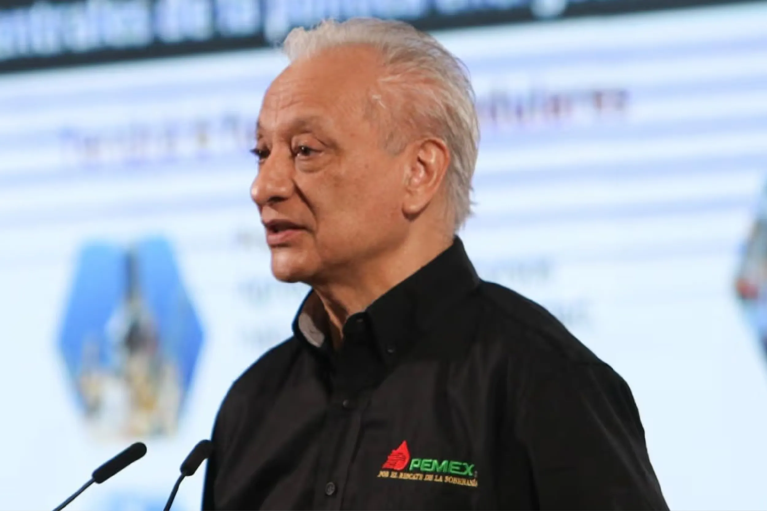 Man in a black shirt with PEMEX logo speaking at a podium with two microphones on a stage.