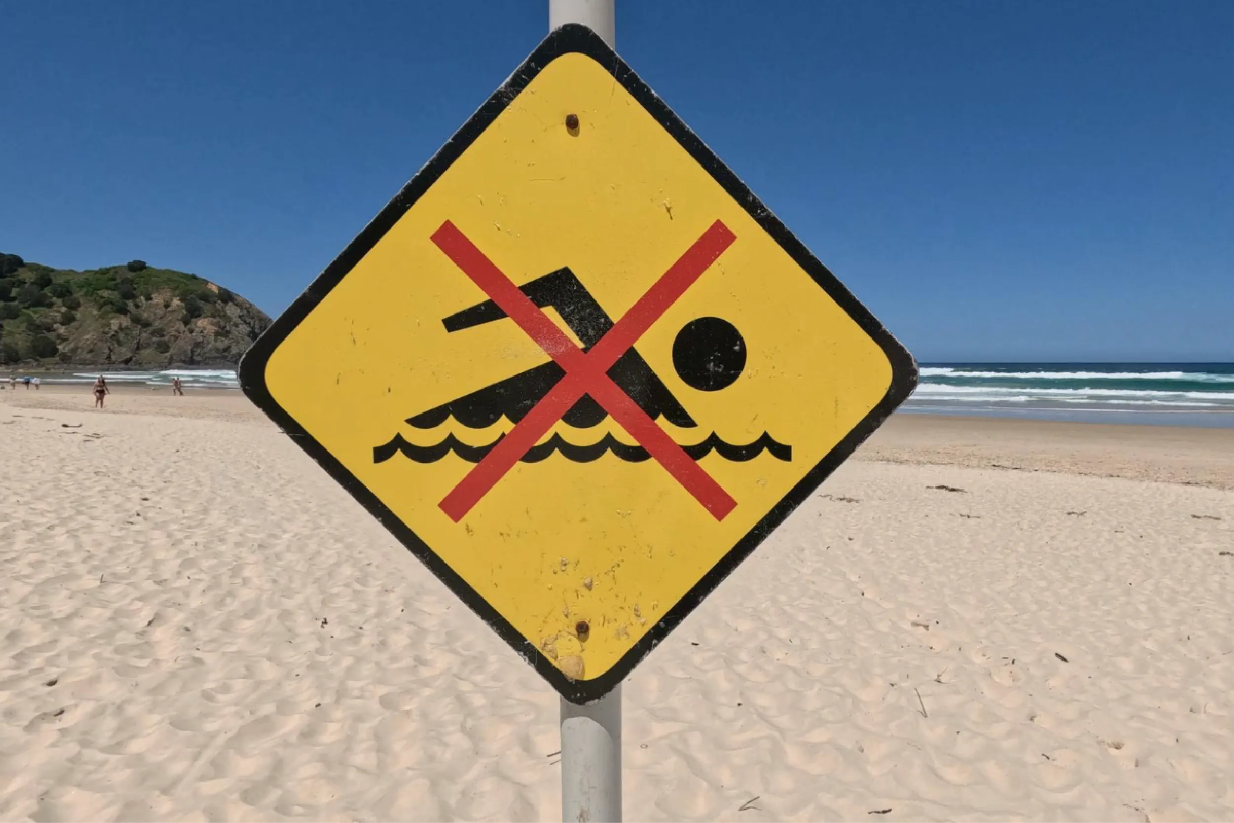 No swimming warning sign on a sandy beach with blue ocean in the background, foreground sign blocks view of water.