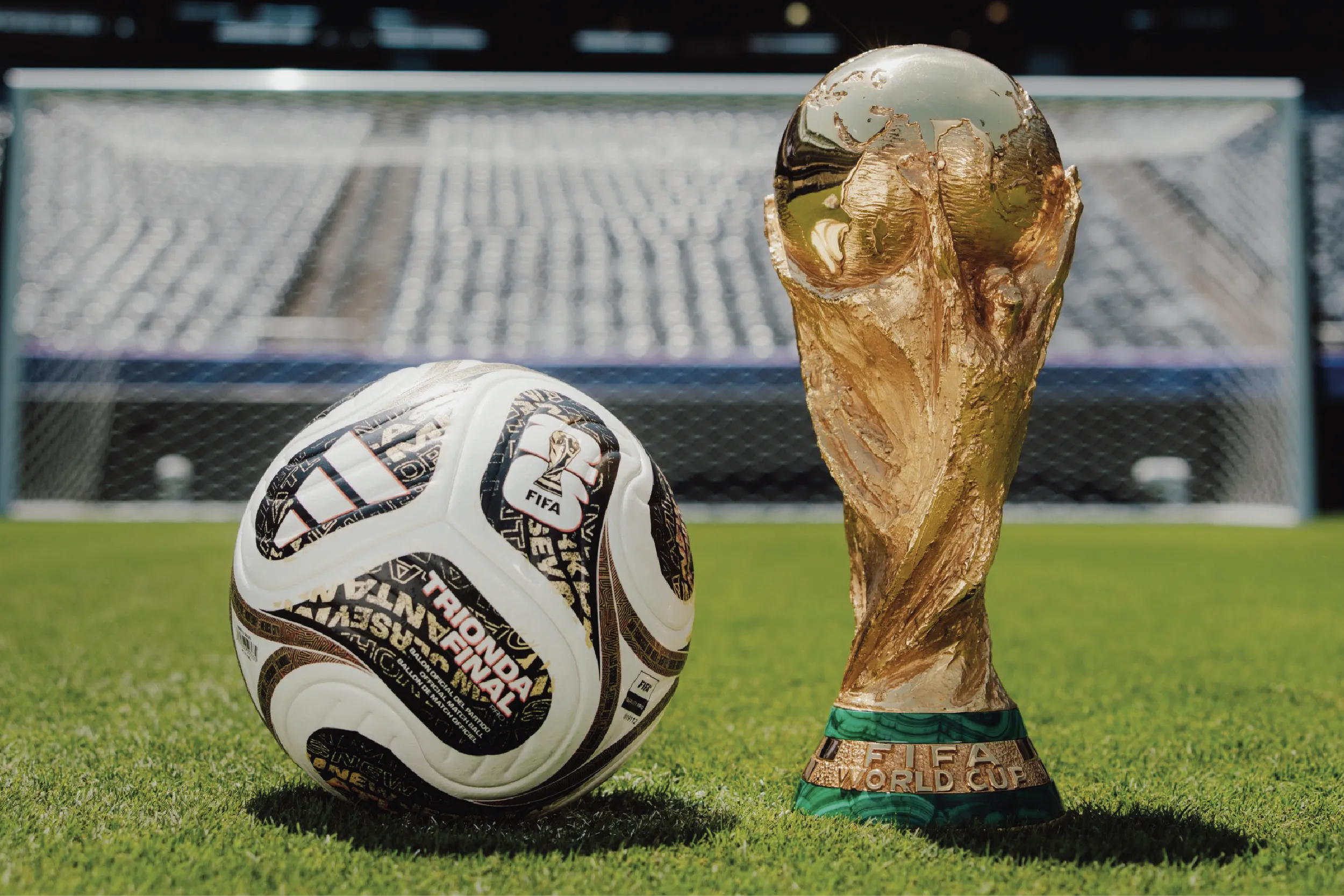 Gold FIFA World Cup trophy beside a patterned soccer ball on a green grass field with a blurred stadium goal in the background.