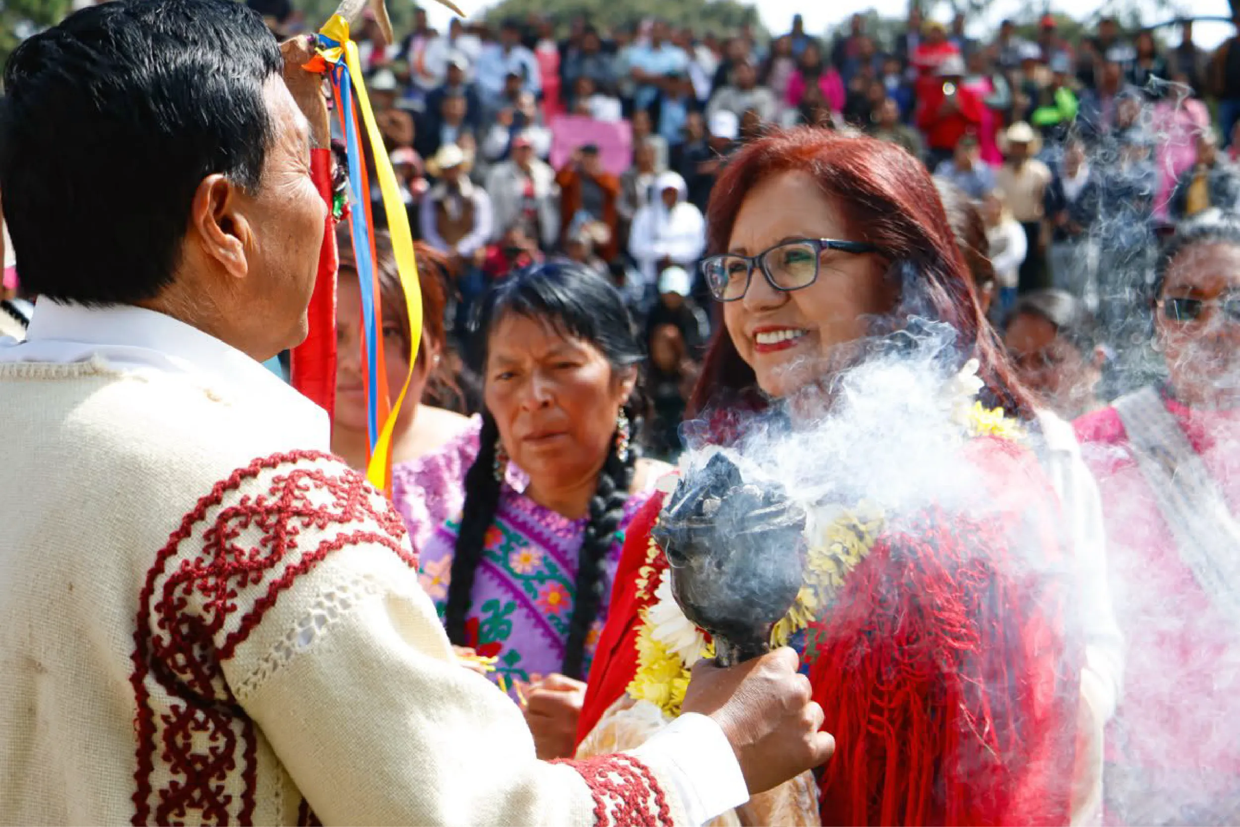 People in bright traditional clothing at an outdoor festival, with ribbons and a smoky foreground, crowds in the background.