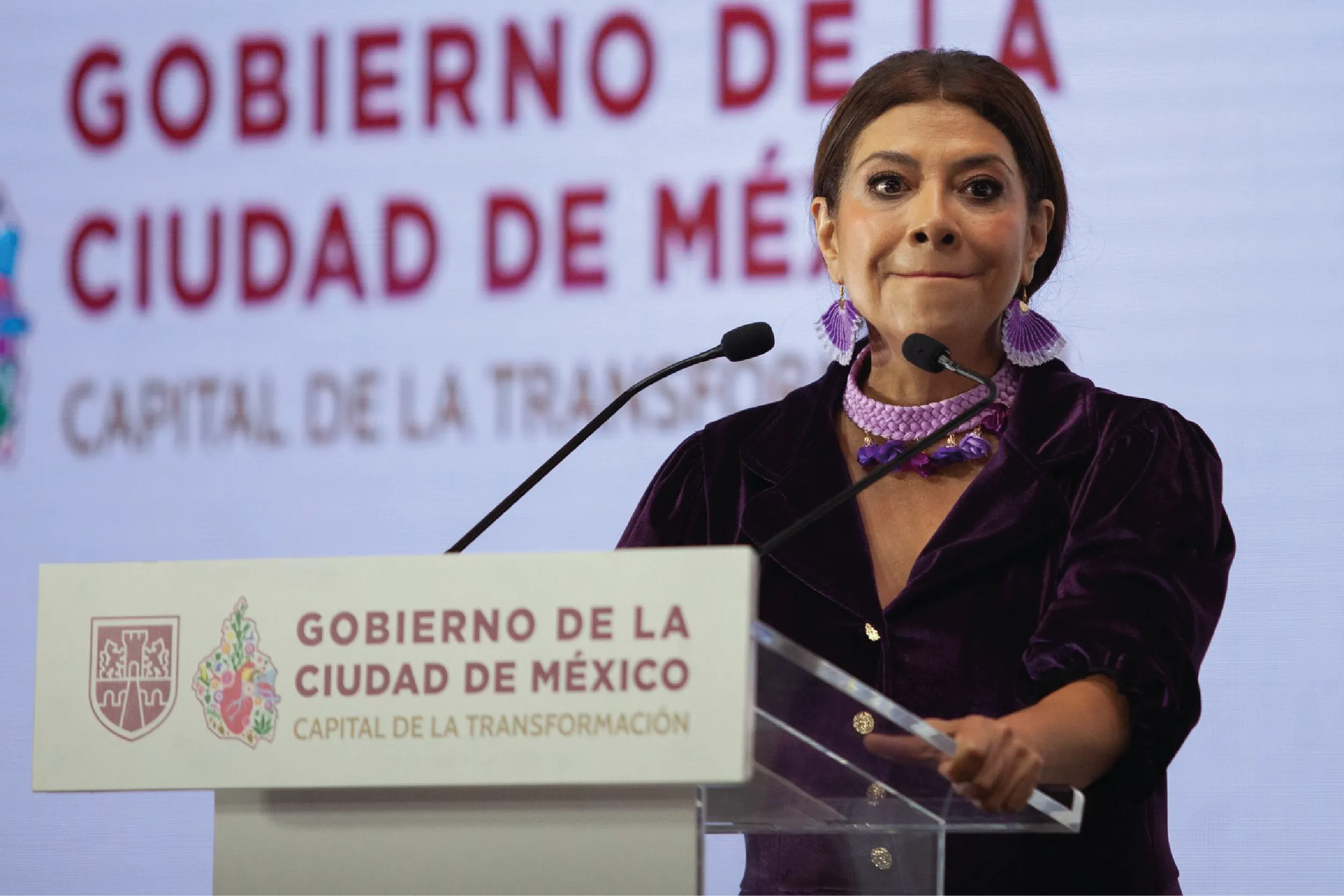 Mujer con vestimenta morada habla en un micrófono detrás de un podio con letreros oficiales de la Ciudad de México.