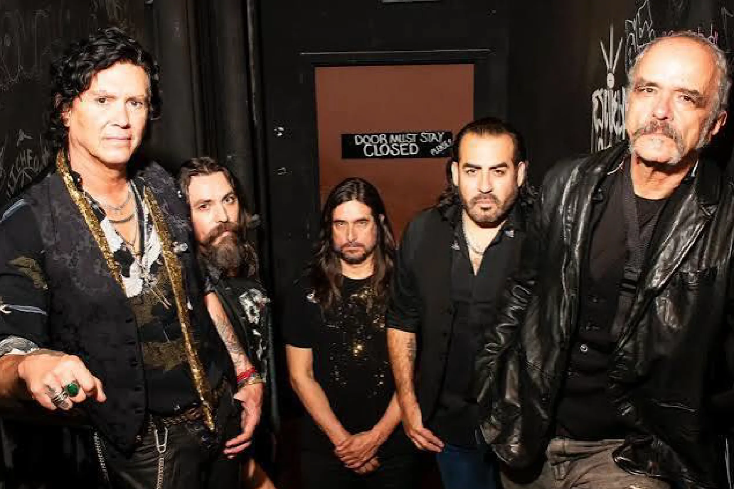 Group of adults in black outfits posing in a backstage hallway with graffiti walls and a closed door in the background, signs visible near the center.