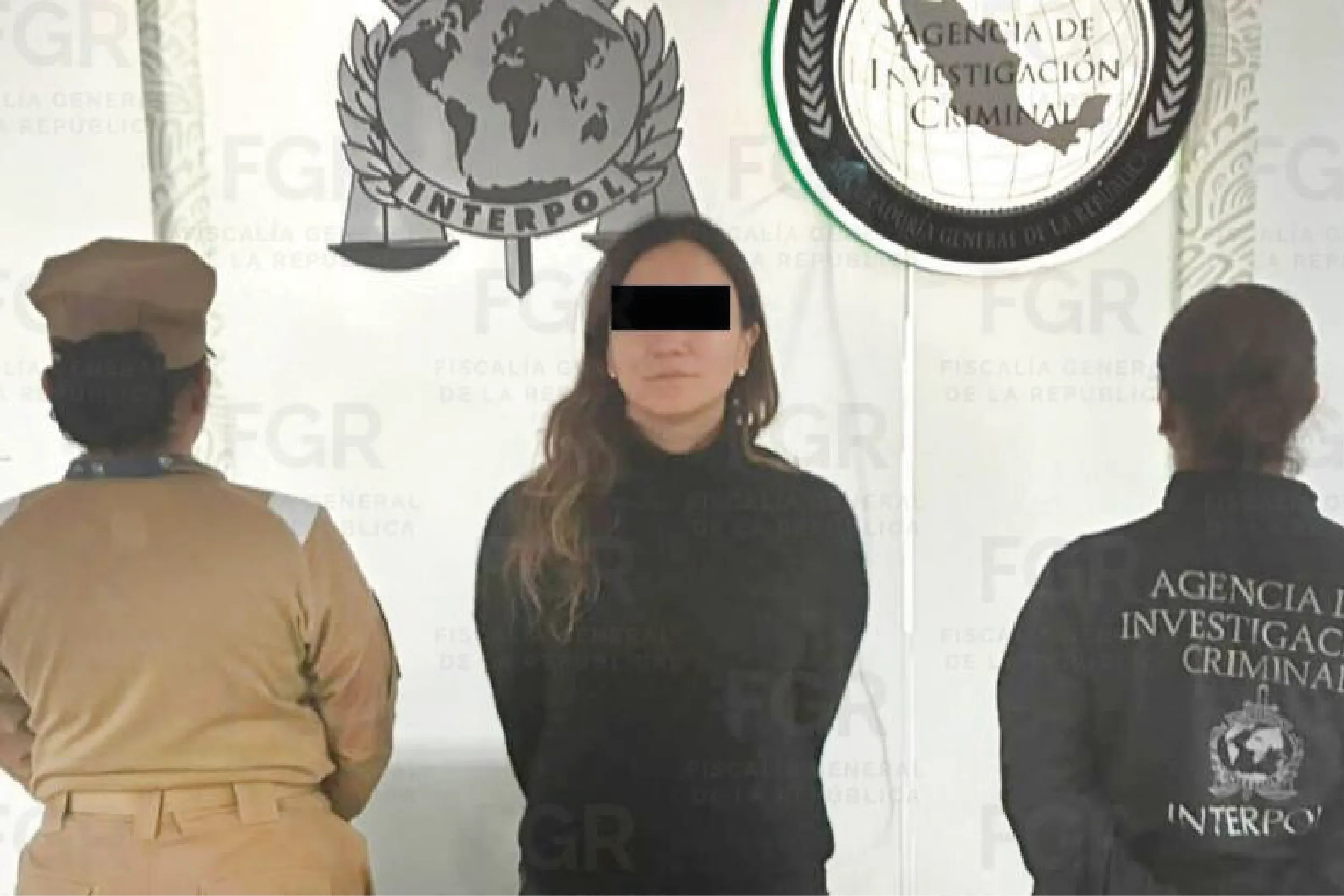 Three people stand in front of a backdrop with government seals; the center person faces the camera with eyes obscured by a black bar.
