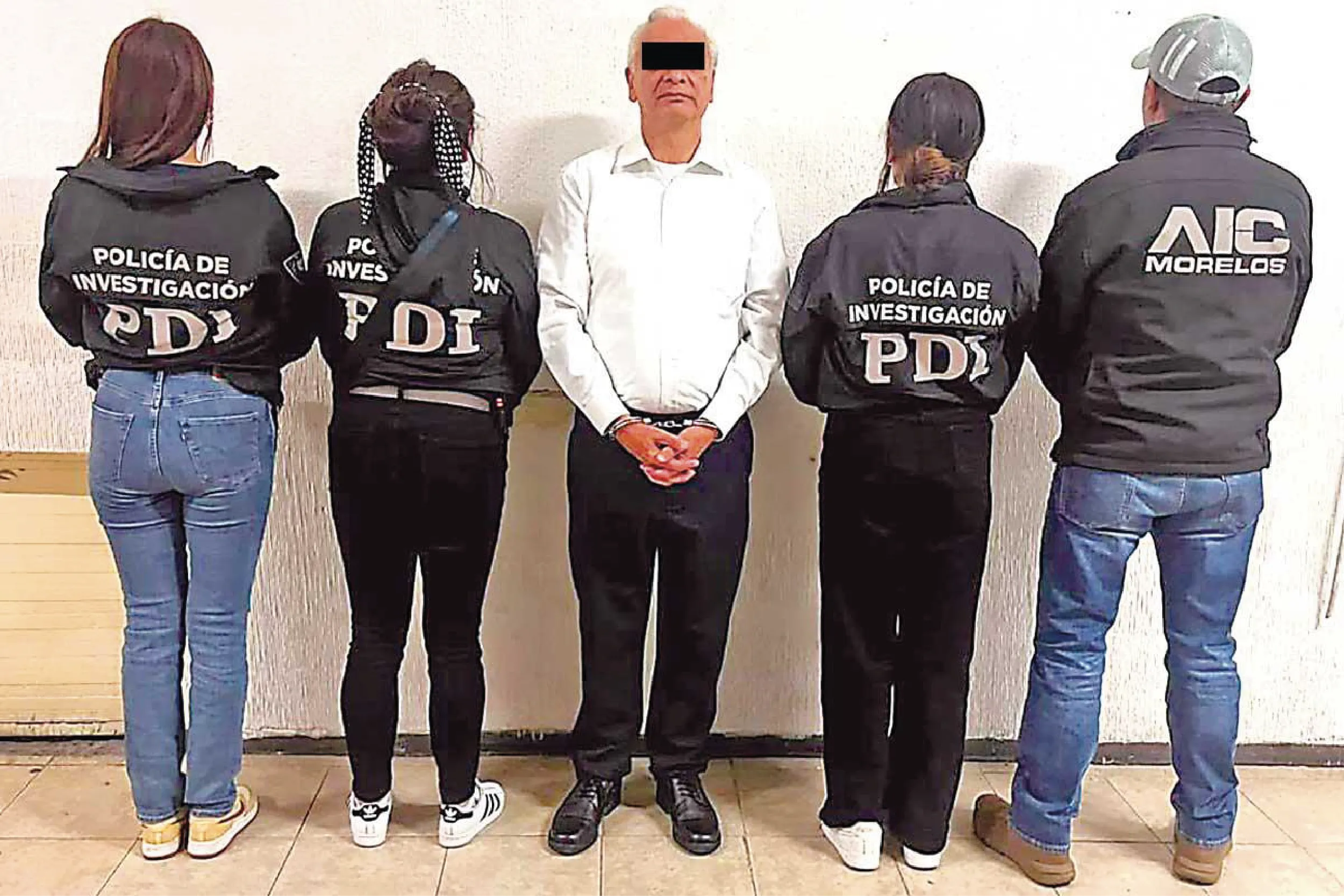Lineup of five people standing against a wall with backs to the camera; four wearing jackets labeled Policía de Investigación (PDI), center man in a white shirt with hands cuffed.