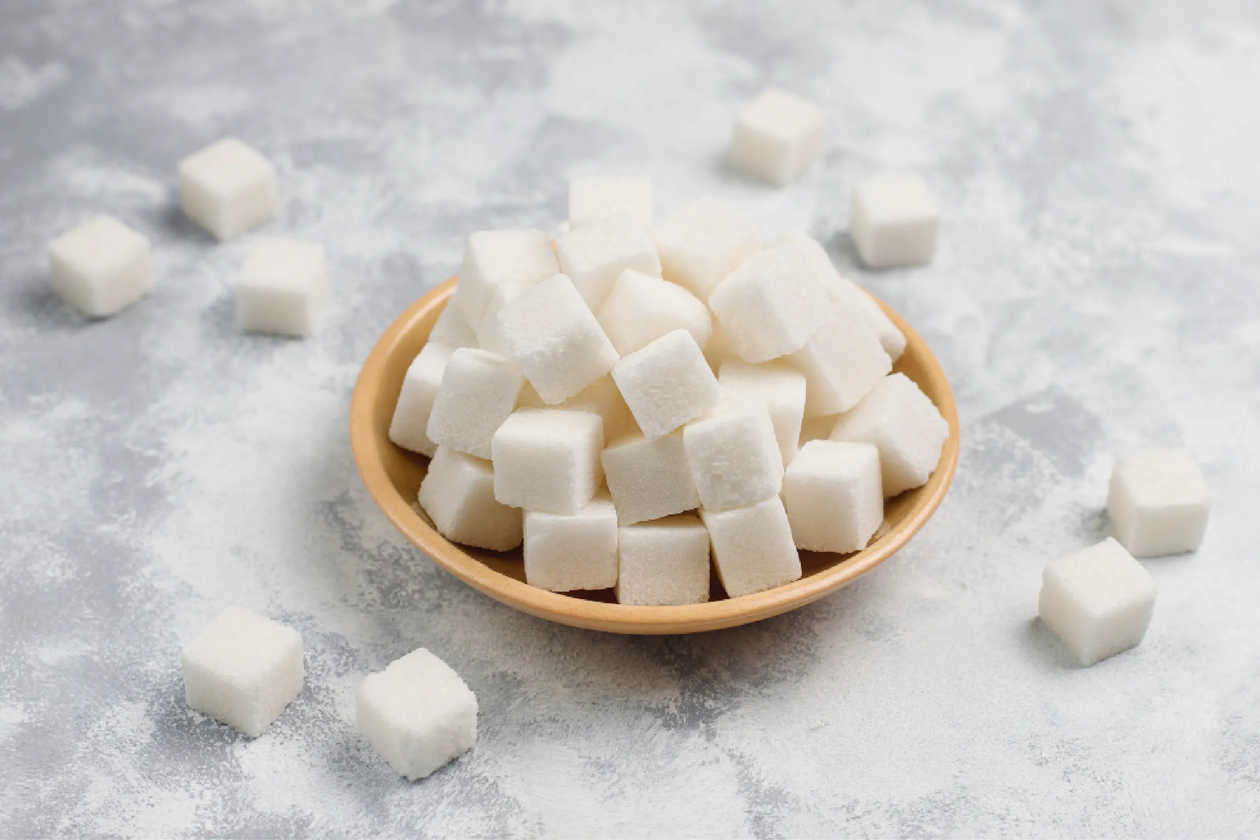 White sugar cubes piled in a wooden bowl on a snowy-gray surface, with cubes scattered around.