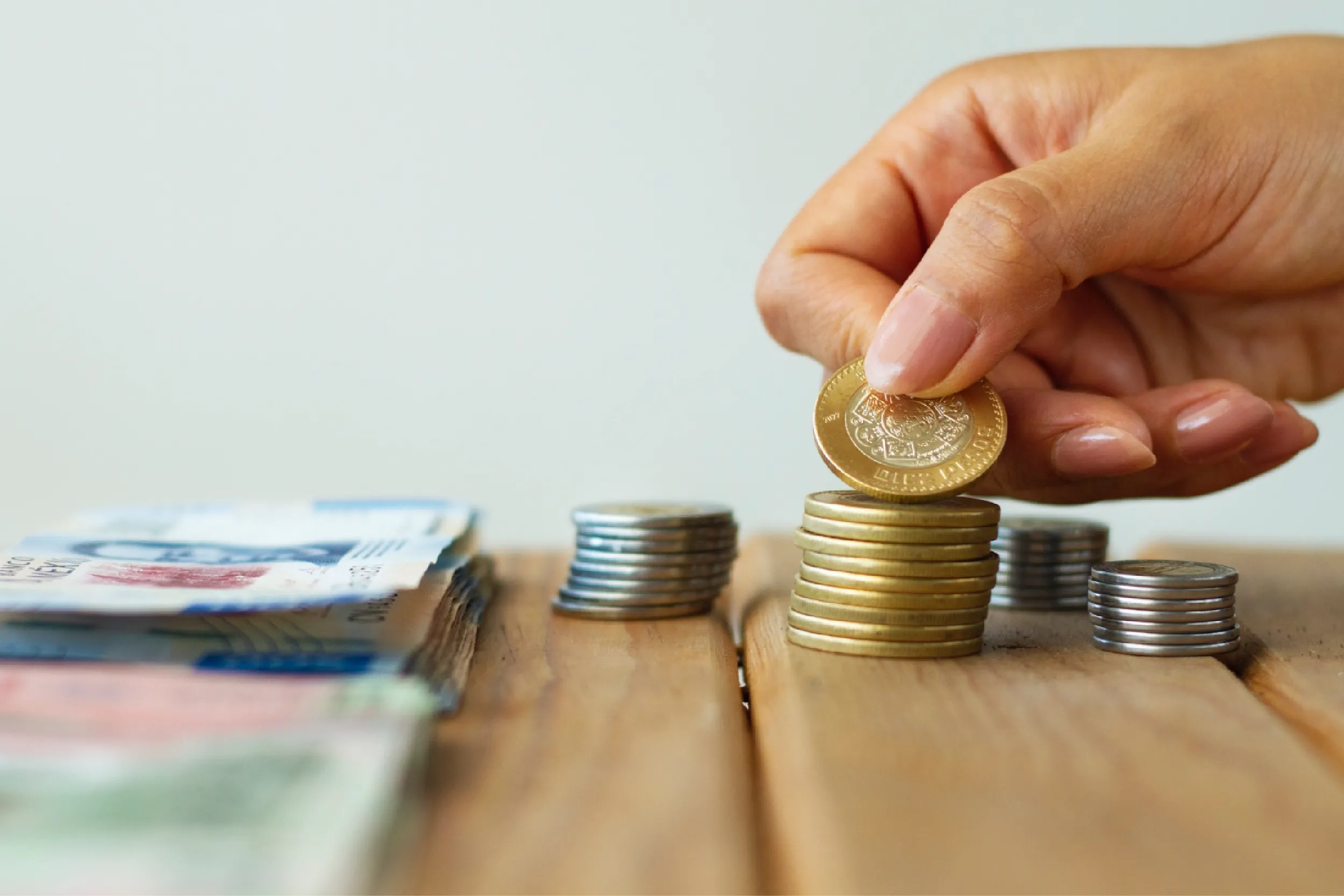 Hand placing a coin on a stack of coins next to scattered banknotes, illustrating saving or budgeting.