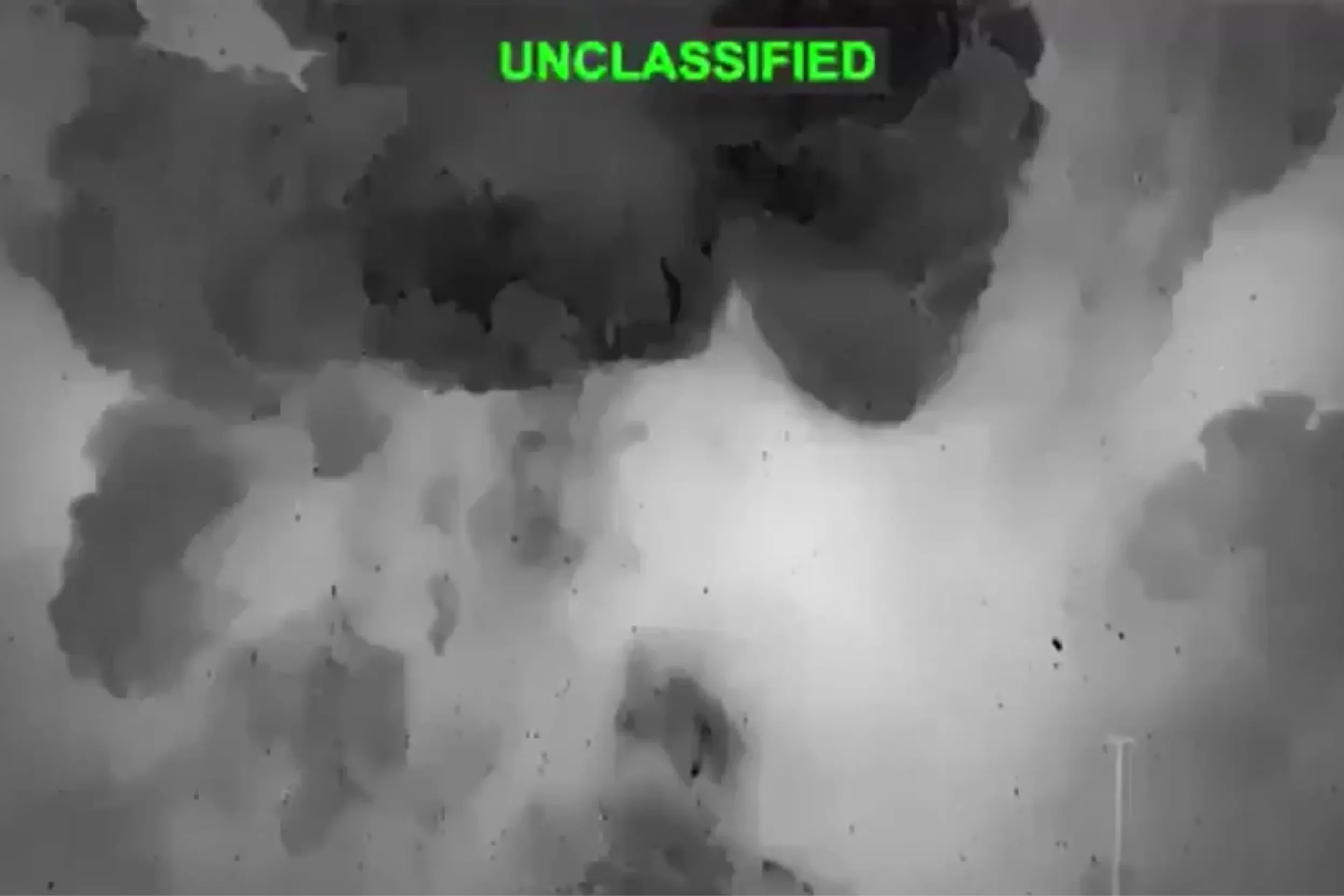 Grayscale surveillance frame showing smoky clouds with a bright green UNCLASSIFIED label at the top.