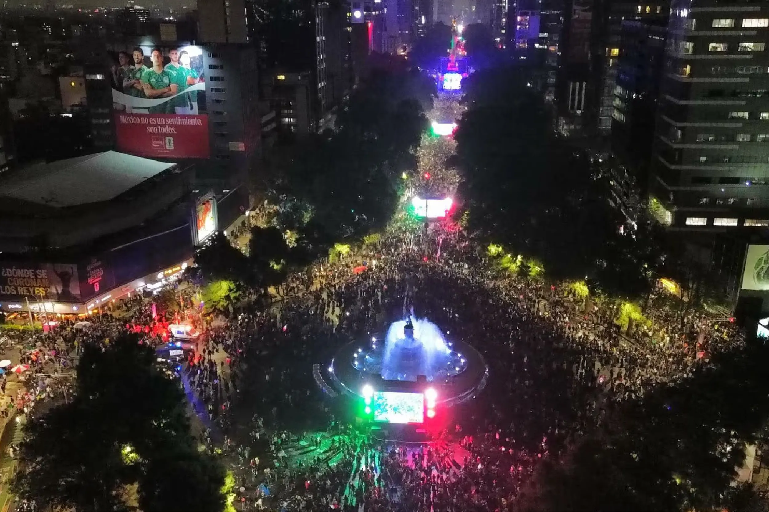 Aerial view of a crowded city square at night, with colorful neon lights and a central illuminated fountain surrounded by buildings.