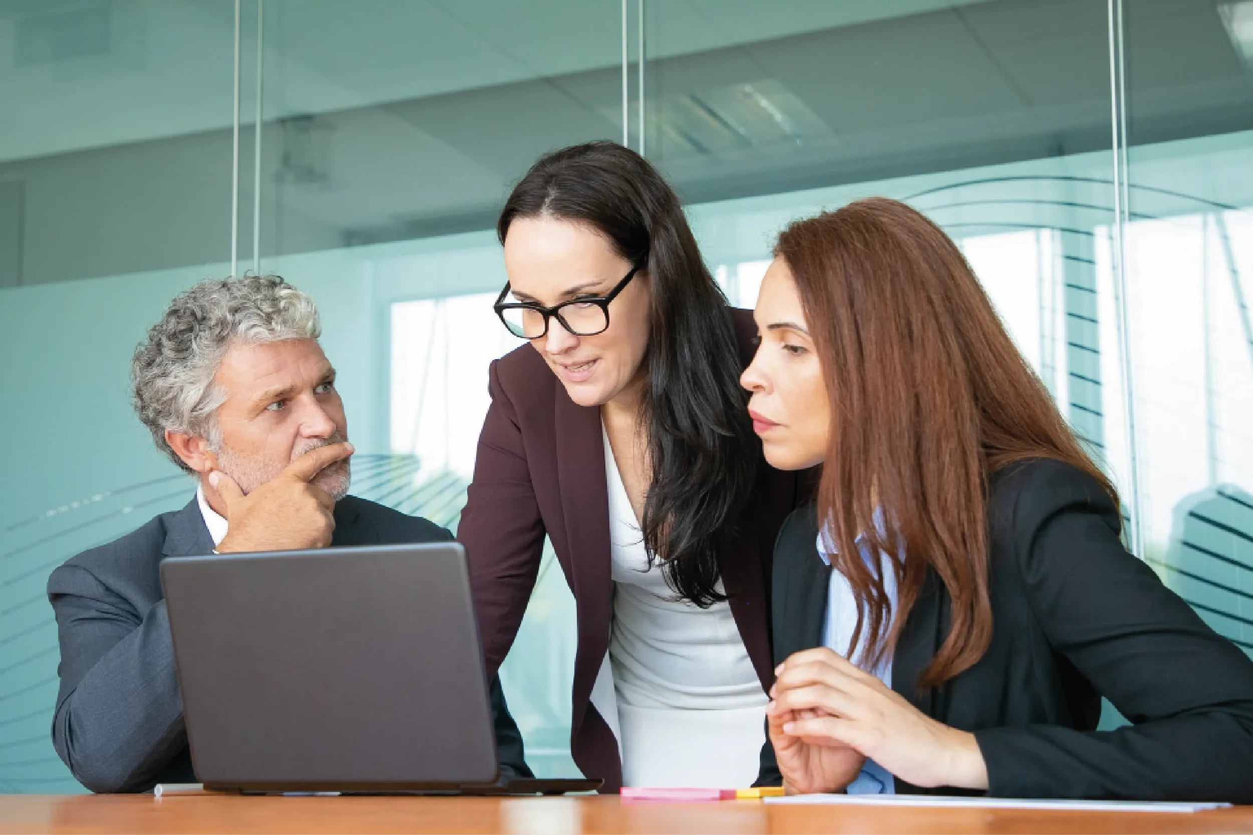 Three business professionals lean over a laptop in a glass-walled conference room, discussing a document.