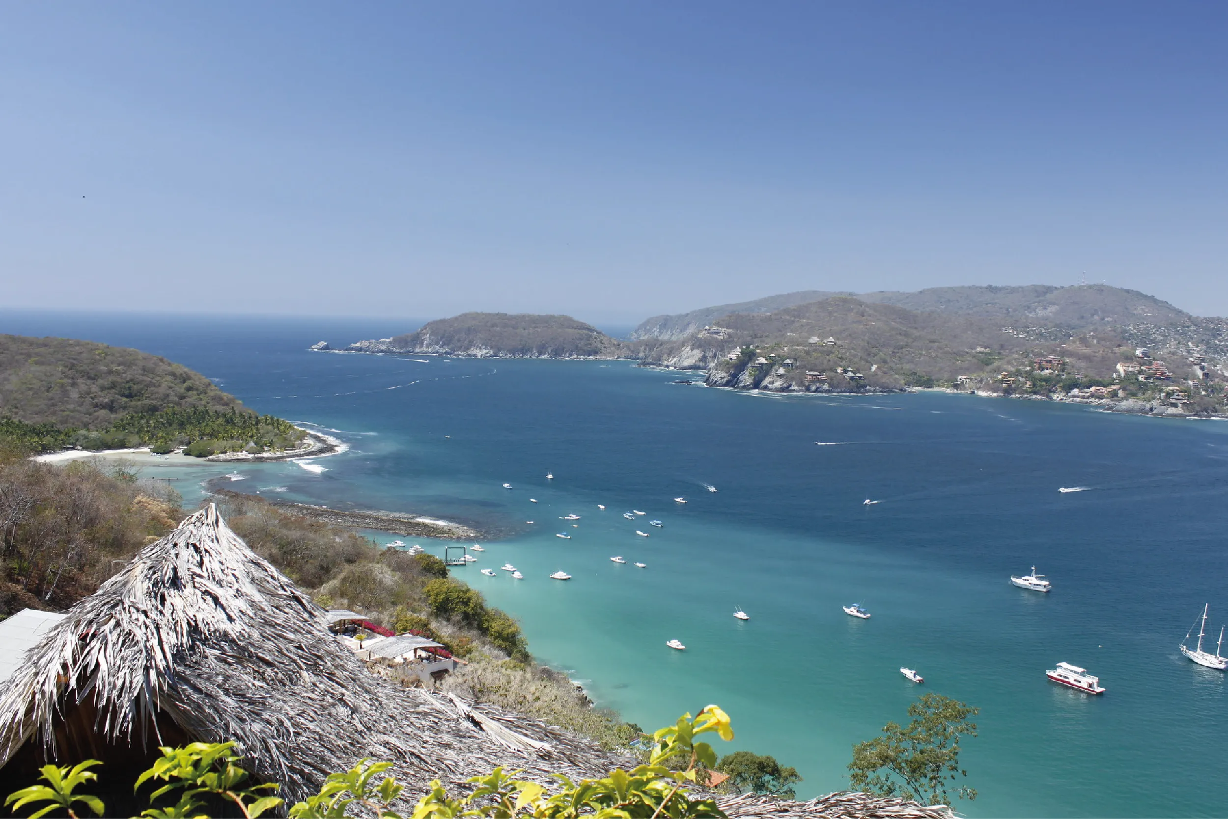 Coastal bay with turquoise water, boats anchored near a rocky shoreline and distant hills under a clear blue sky.