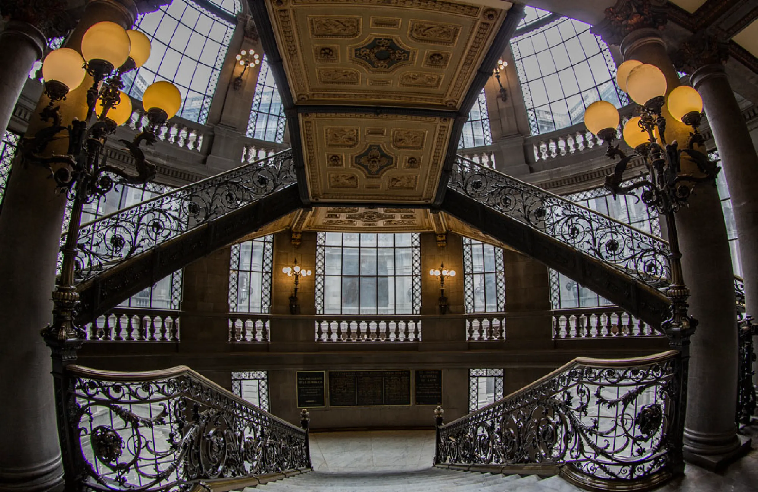 Grand interior staircase with ornate wrought-iron railings, stone columns, and warm globe lights in a historic lobby.