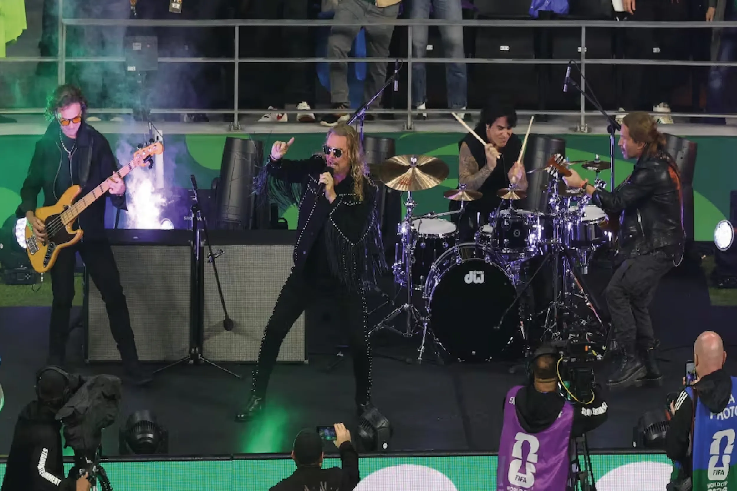 Rock band performing on stage with guitarist left, singer center, drummer, and another guitarist on right amid colorful lights and smoke.