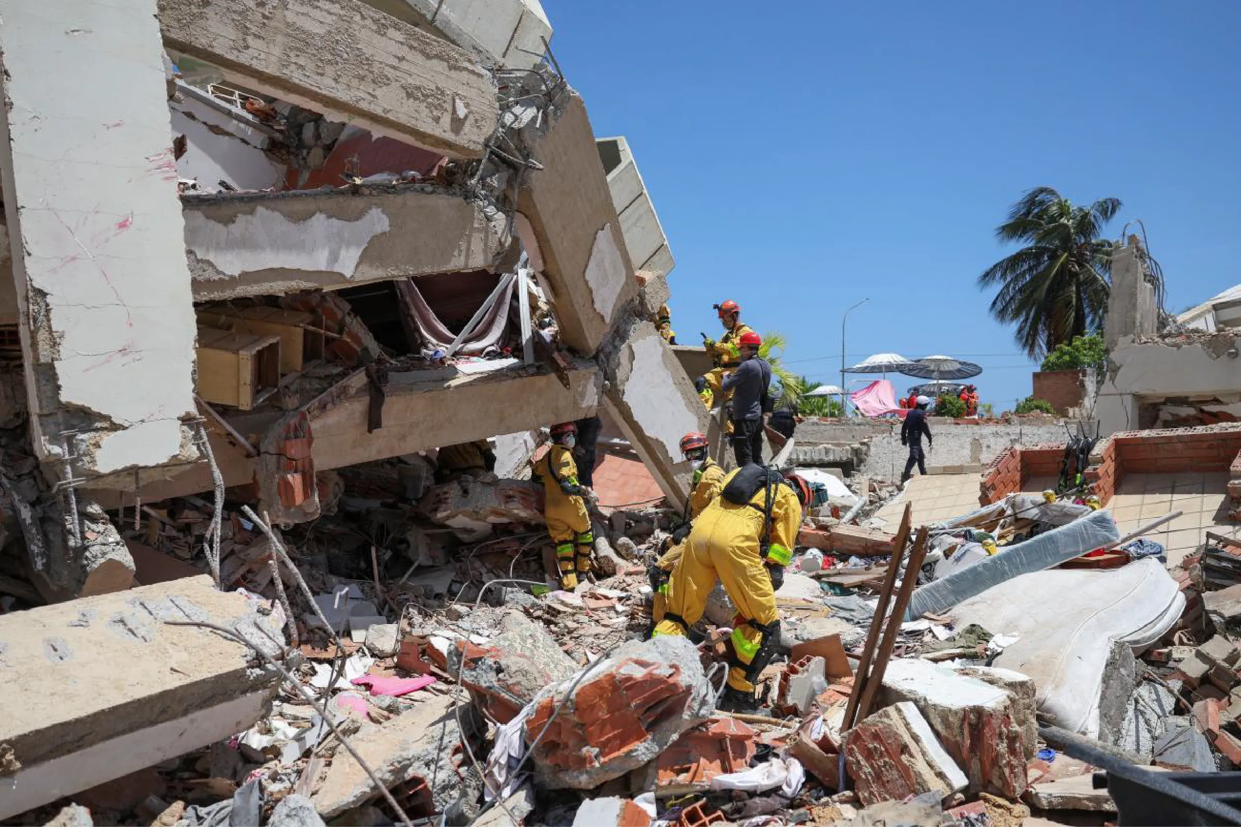 Firefighters in yellow gear search through rubble of a collapsed building under a clear blue sky.