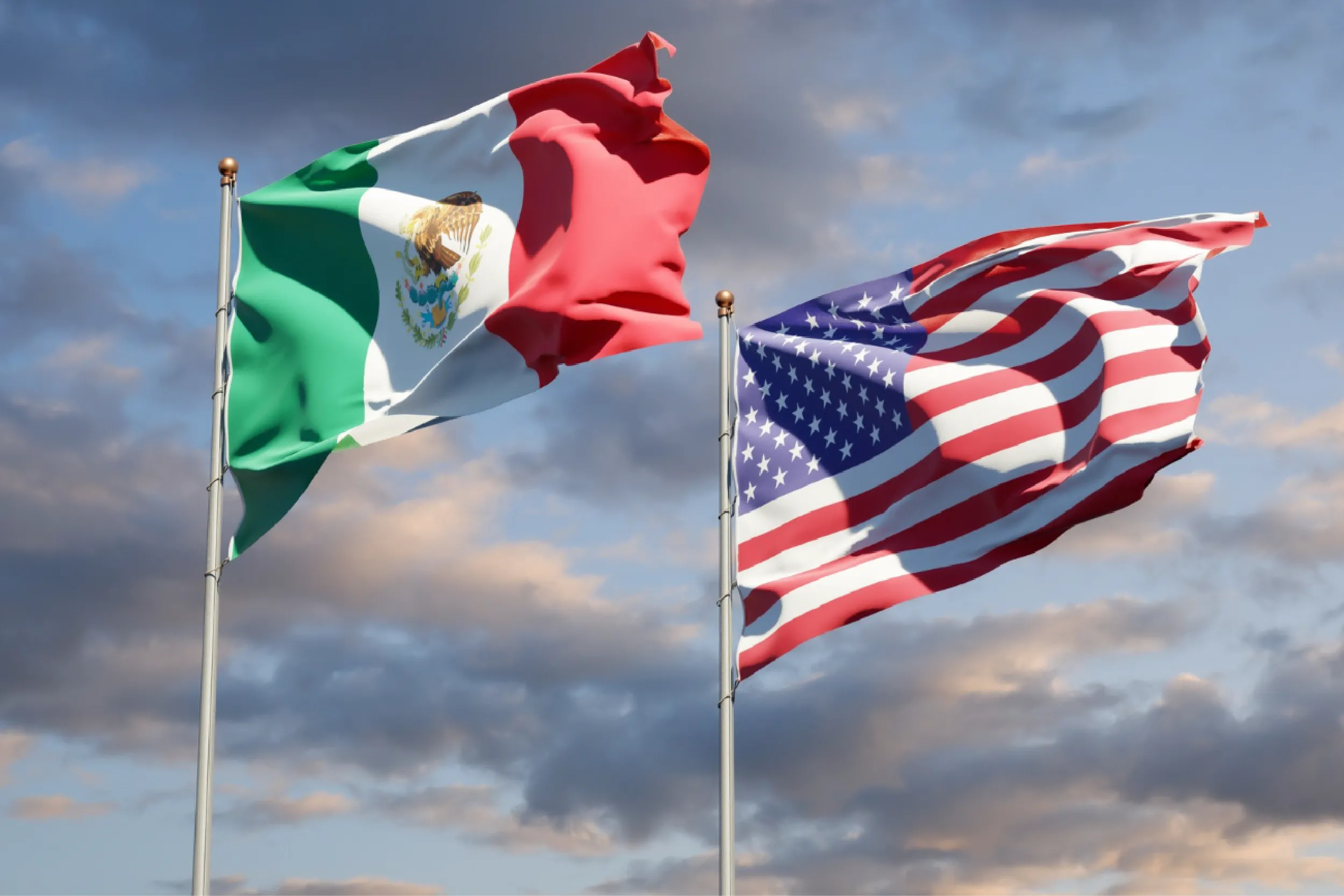 Mexican and American flags waving on flagpoles against a cloudy blue sky.