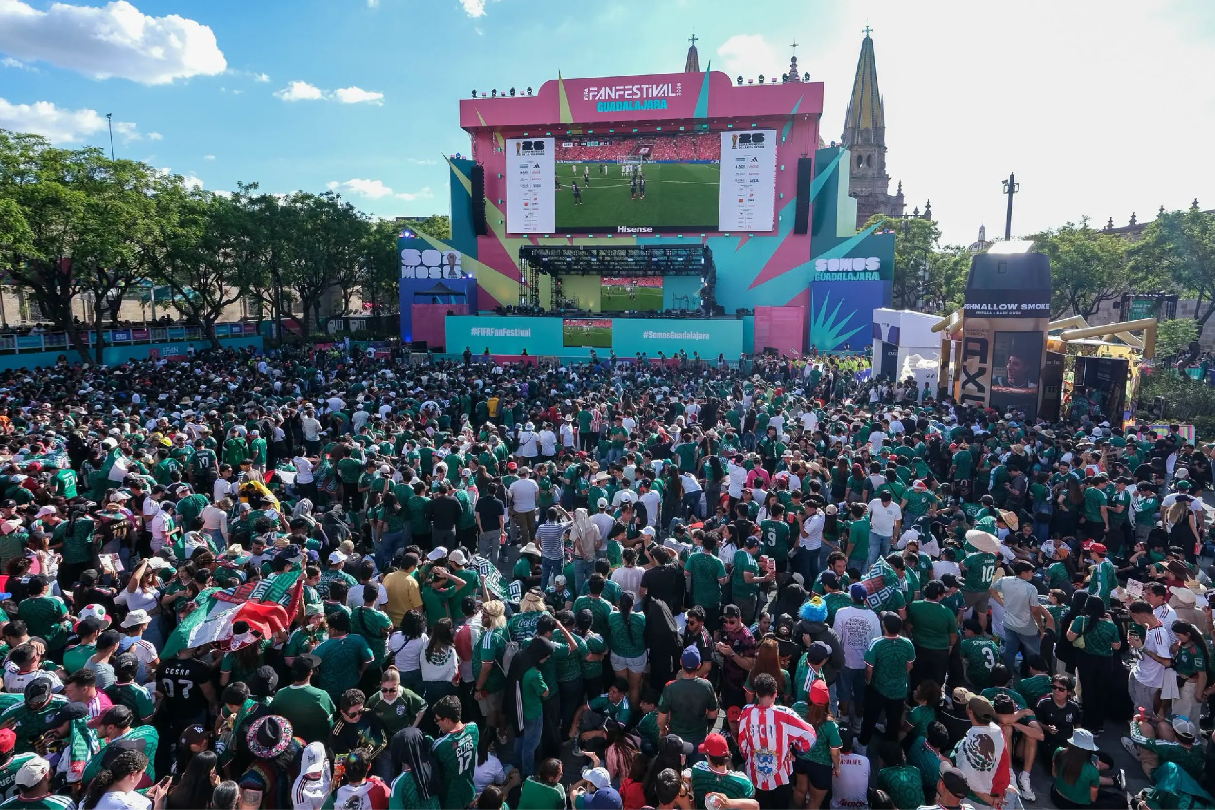 Thousands of fans in green jerseys fill an outdoor festival venue, watching a soccer match on a large colorful stage screen (FIFA Fan Festival Guadalajara).