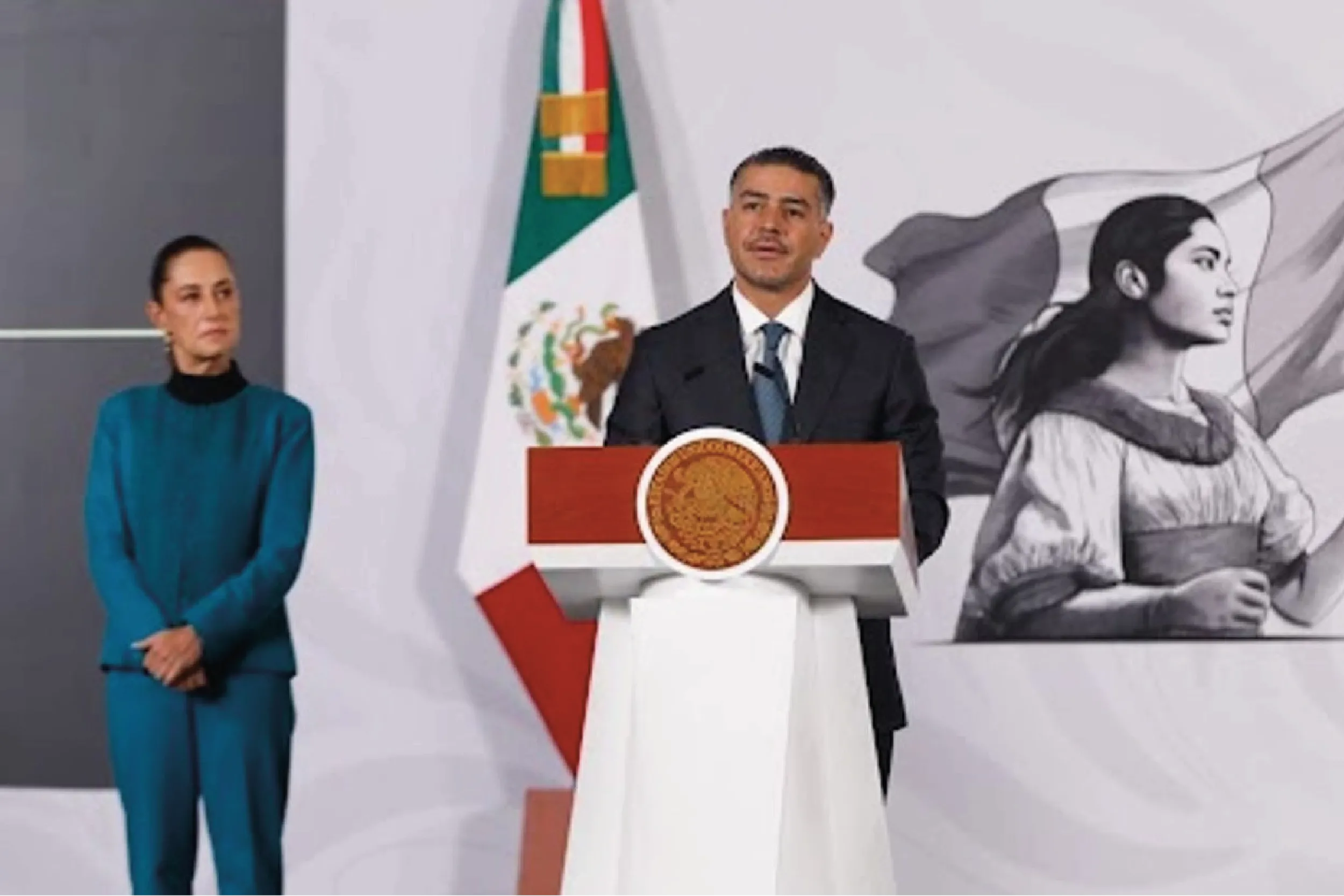 Man in a dark suit speaking at a podium with the Mexican flag behind; a woman in teal stands to the left.