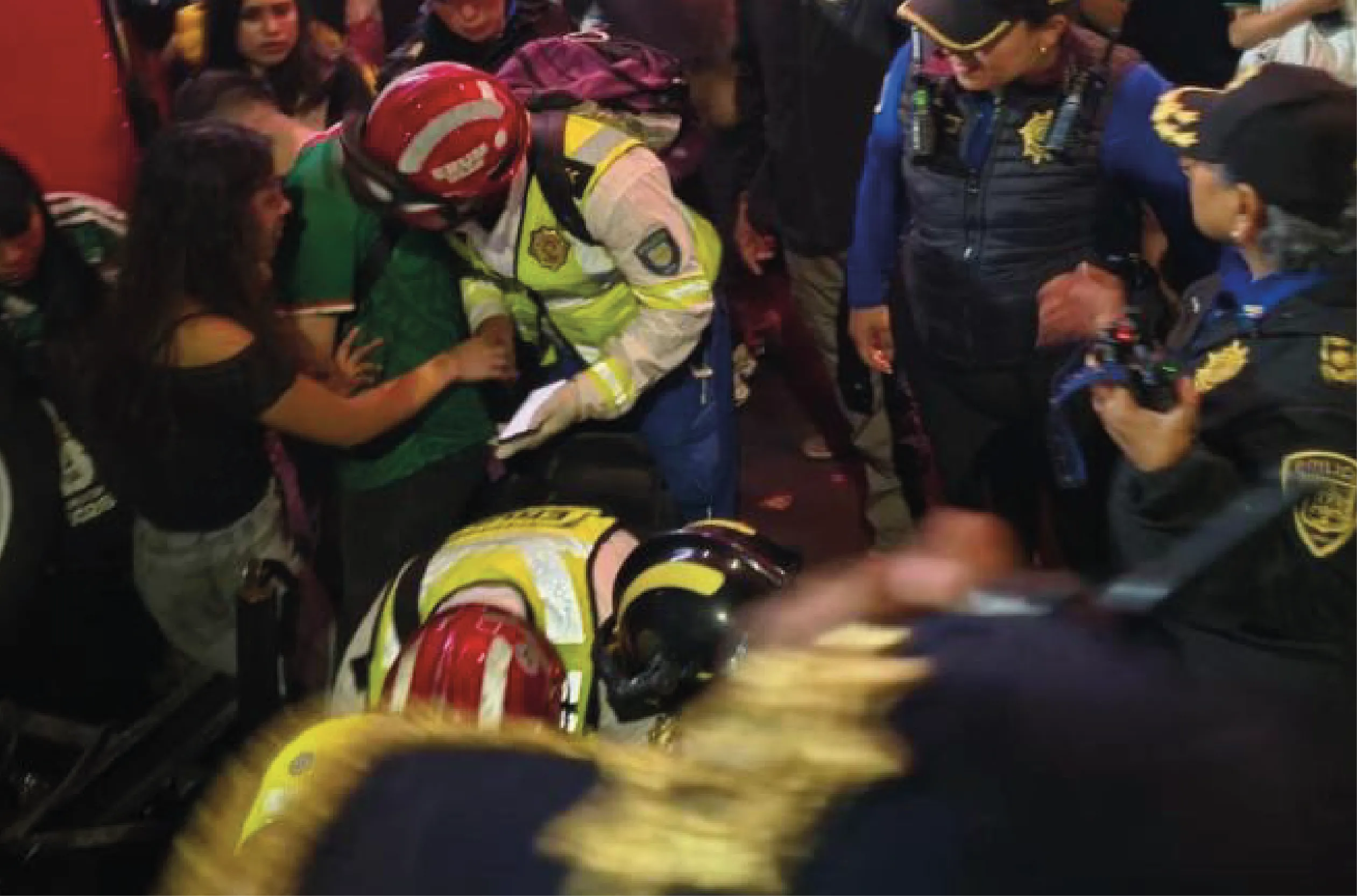 Crowded indoor scene with motorcycle riders in helmets and neon jackets assisting someone on the floor.
