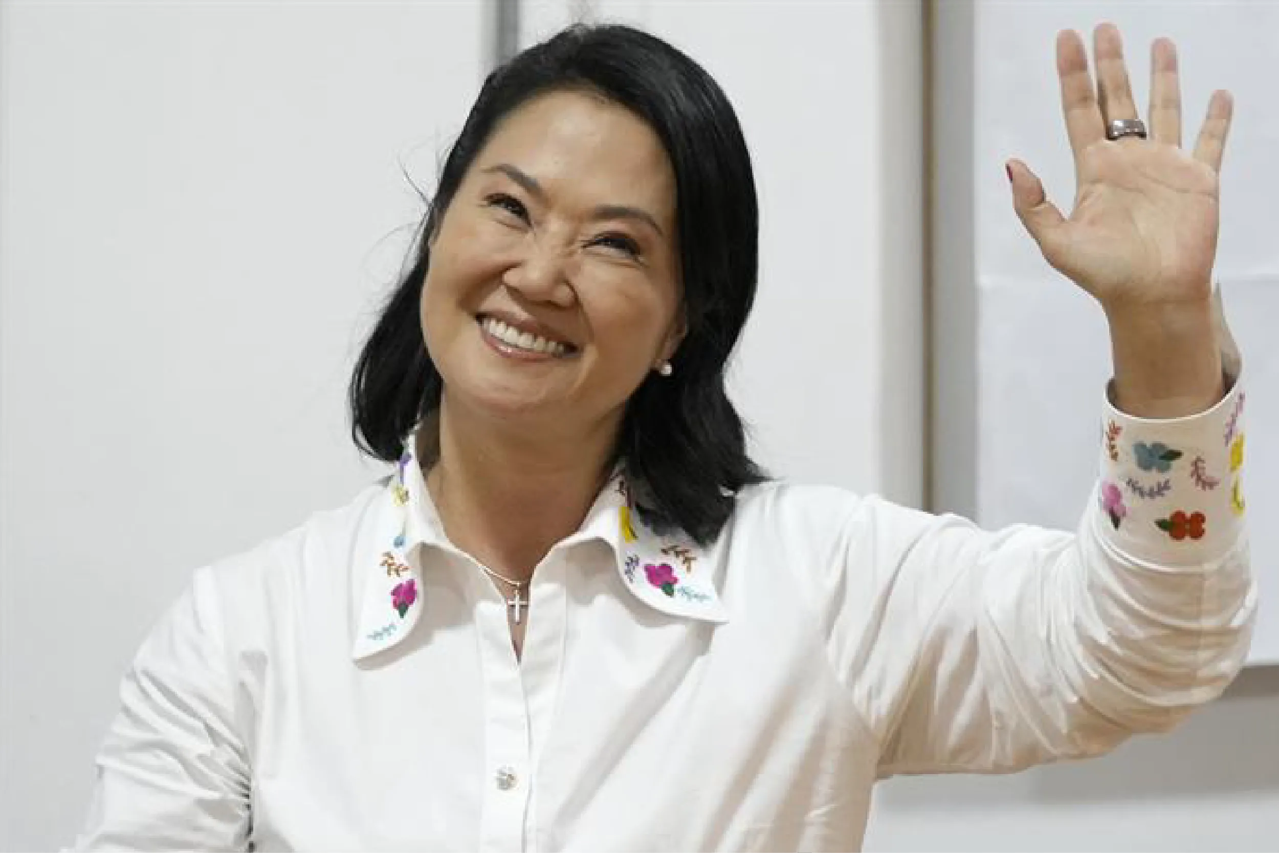 Smiling woman in a white shirt raises her right hand with a ring visible on her ring finger, wearing a colorful butterfly-patterned cuffed sleeve.