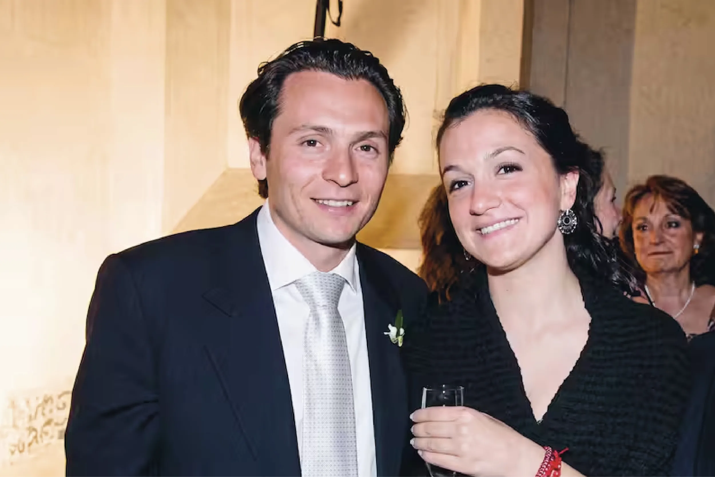 Smiling couple in formal attire posing for a photo at a reception; man in a dark suit and light tie, woman in a black dress with earrings, holding a champagne flute, guests in the background