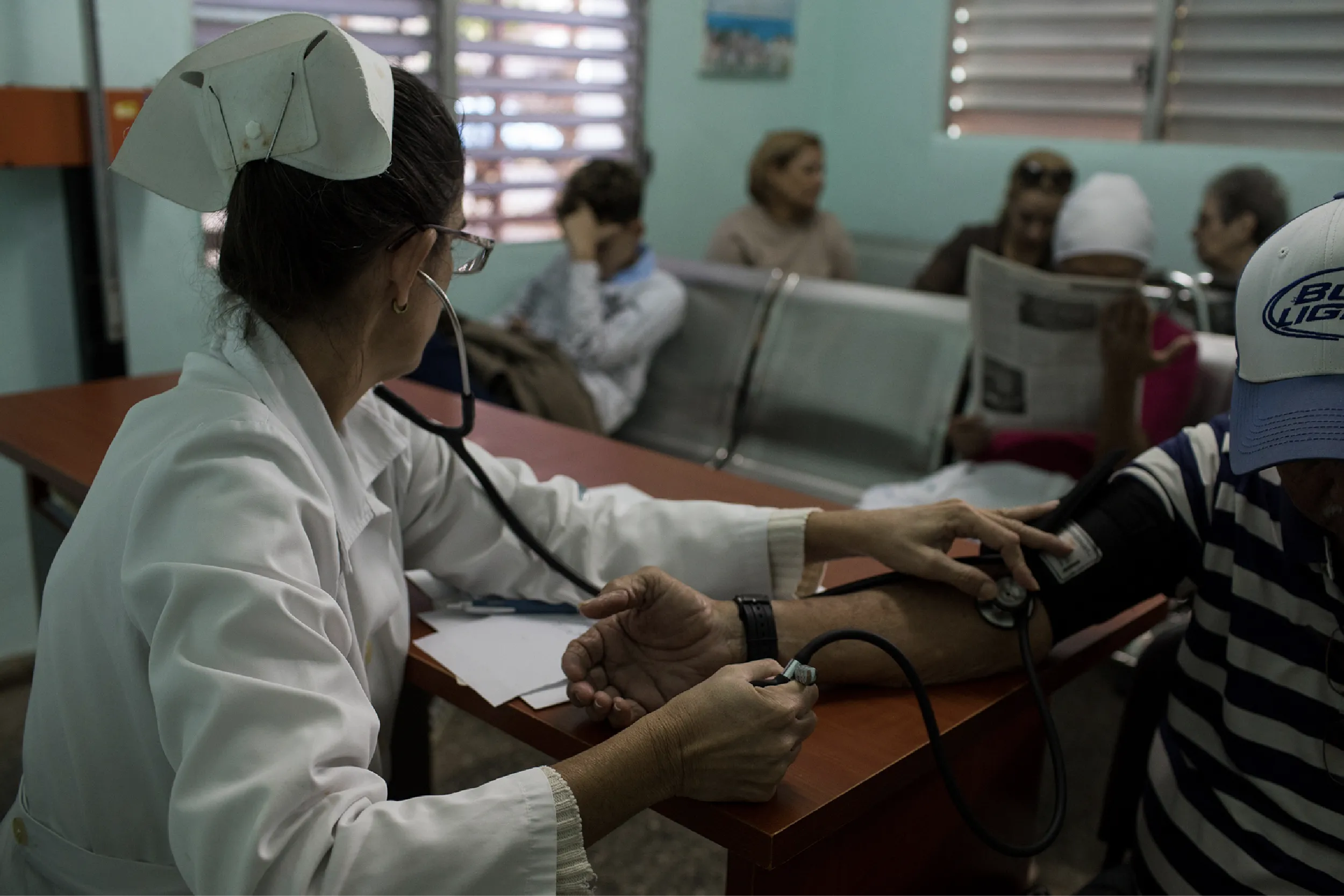 Nurse in a white coat takes a patient’s blood pressure with a cuff at a clinic desk, with other people waiting in the background.
