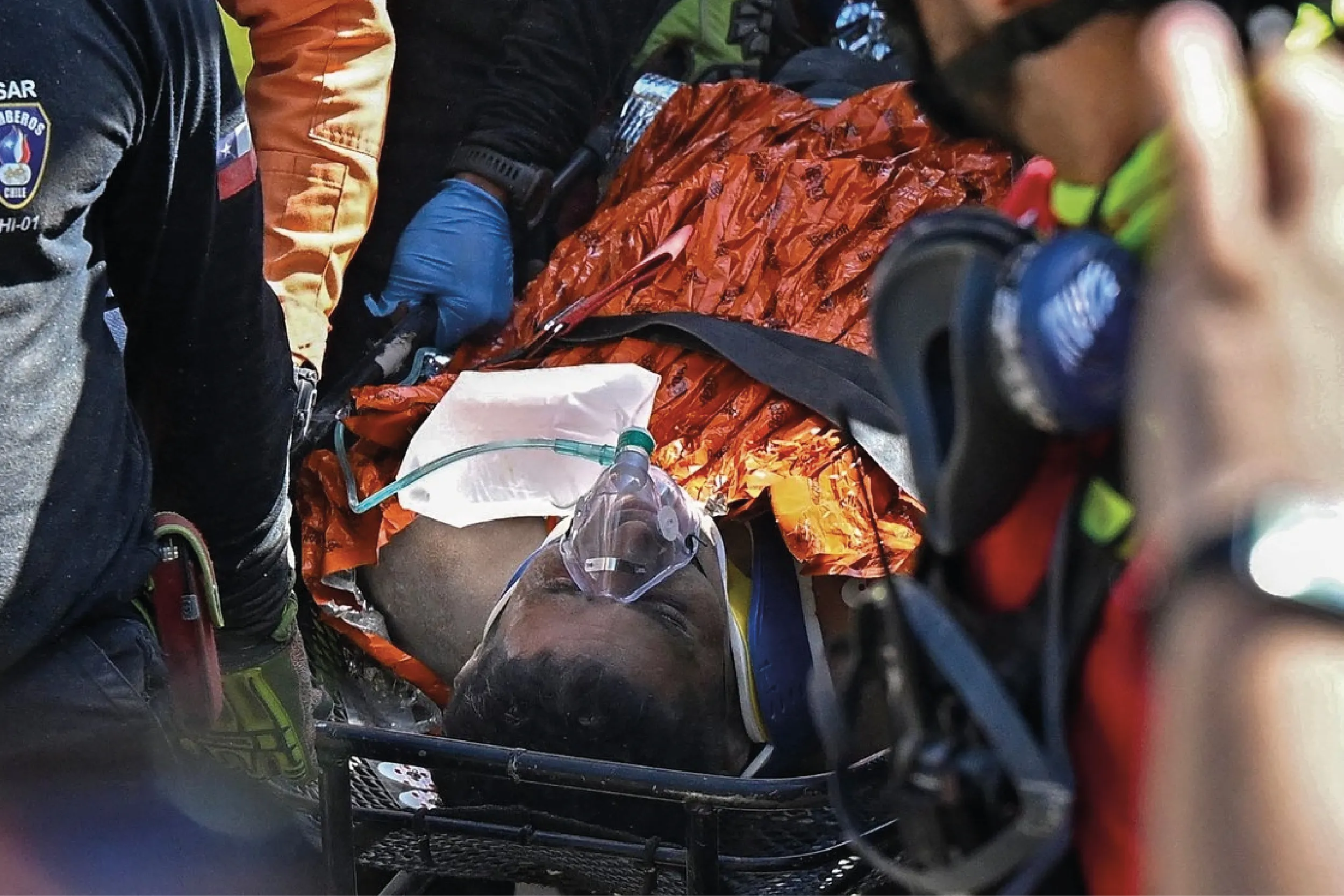 Emergency responders in gloves and dark gear attend to a patient on a stretcher, with an orange rescue bag and an oxygen mask nearby.