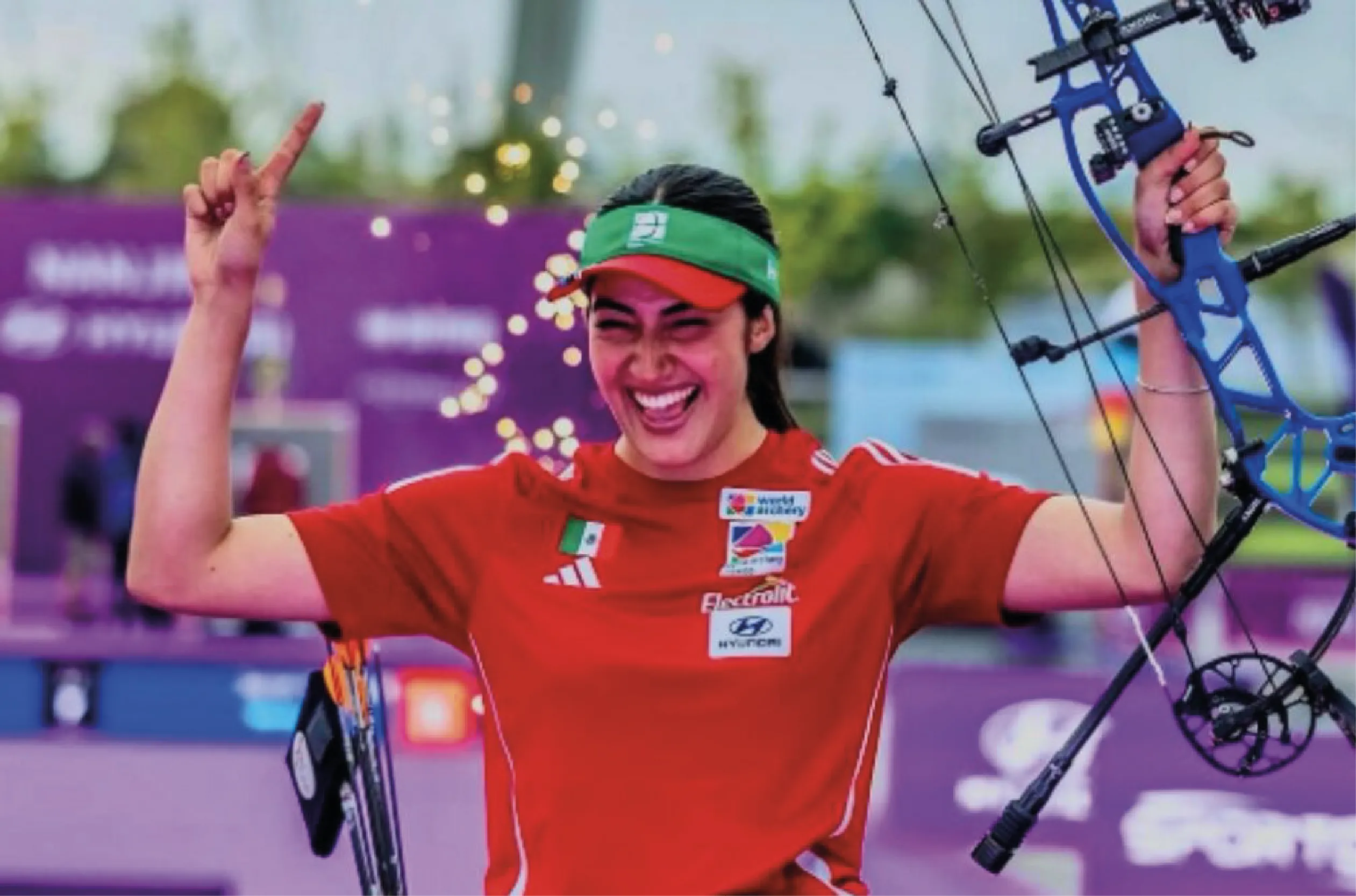 Female archer in a red shirt and green headband celebrates, raising a blue compound bow in the air at a competition venue.