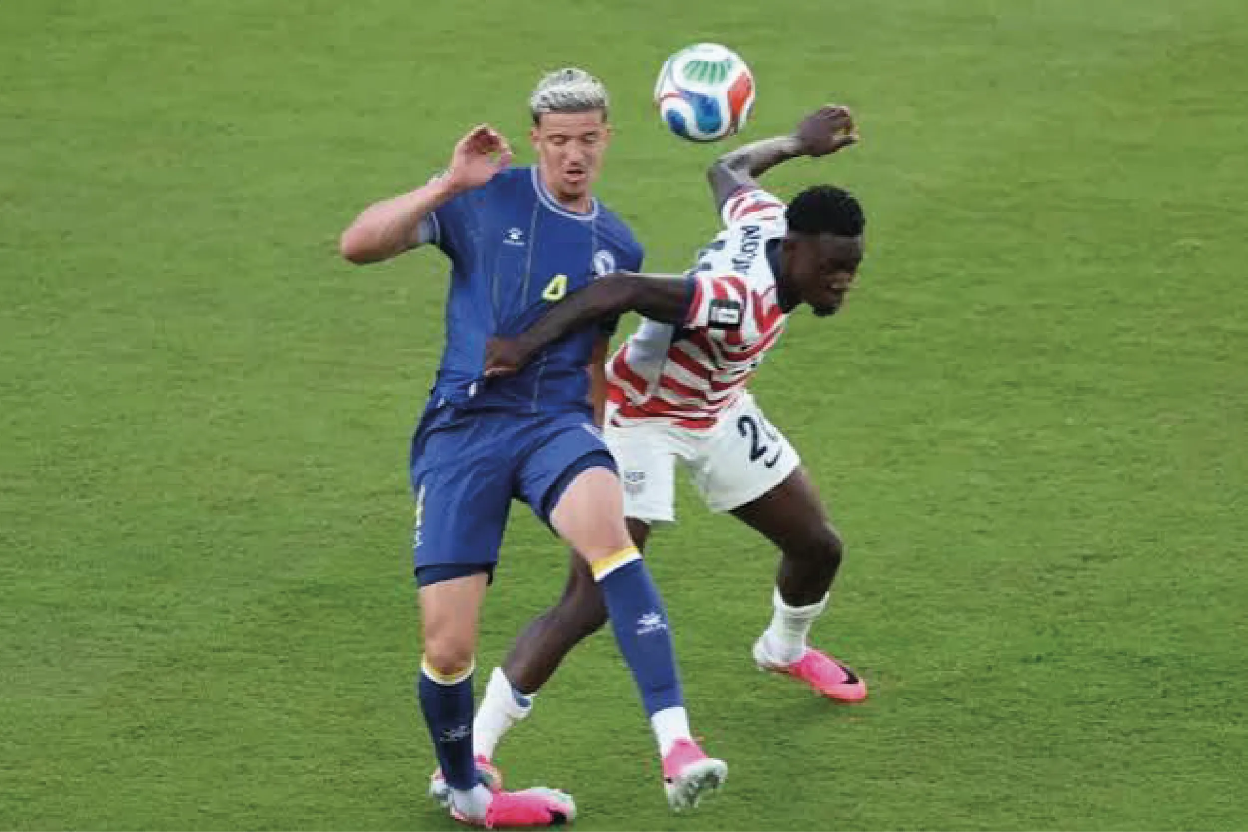 Two soccer players chase a ball in midair during a match; one in blue, the other in red-and-white stripes, on green turf.