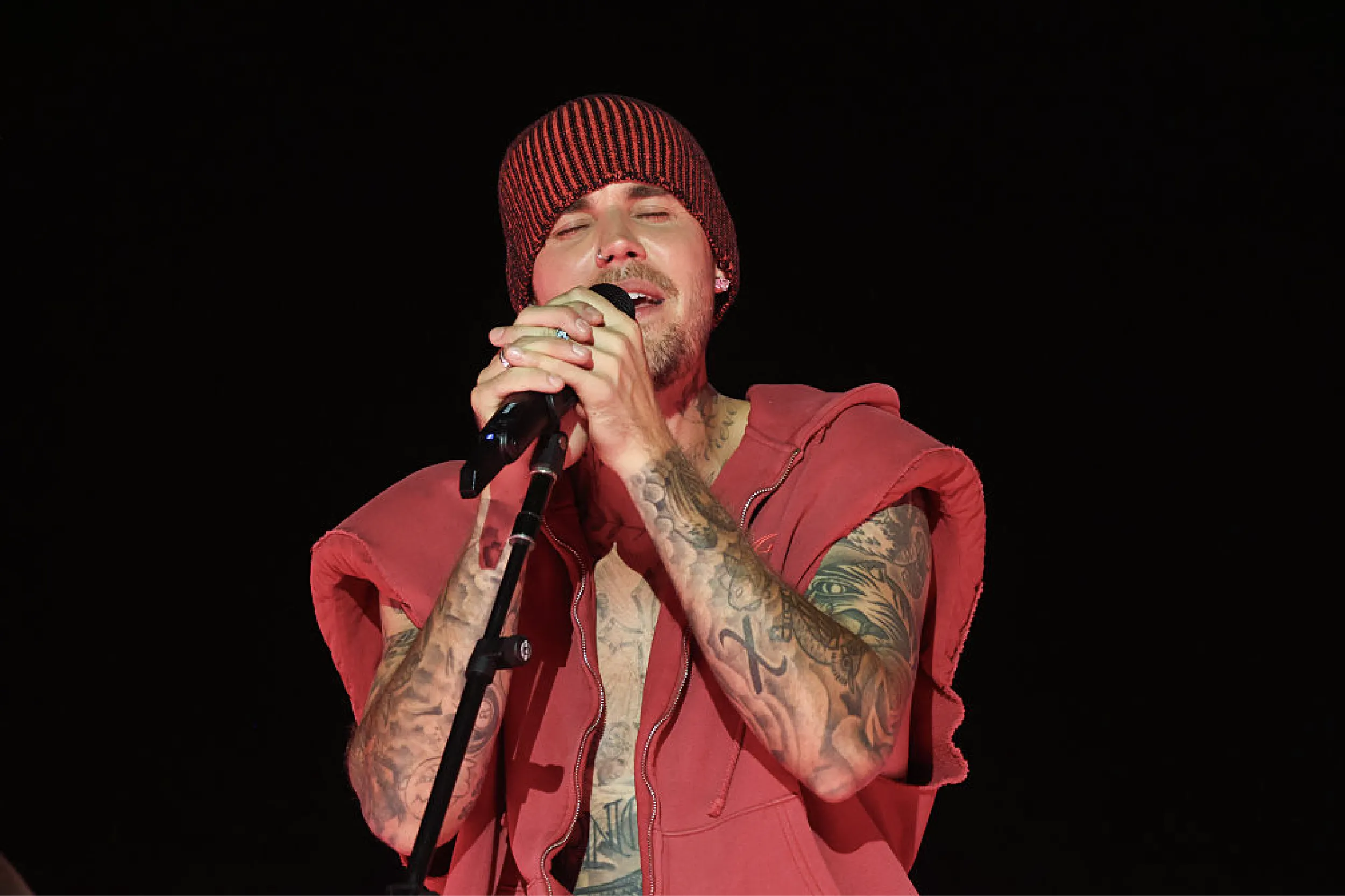 Male singer wearing a red knit beanie and sleeveless red hoodie, singing into a microphone on a dark stage, arms covered in tattoos.