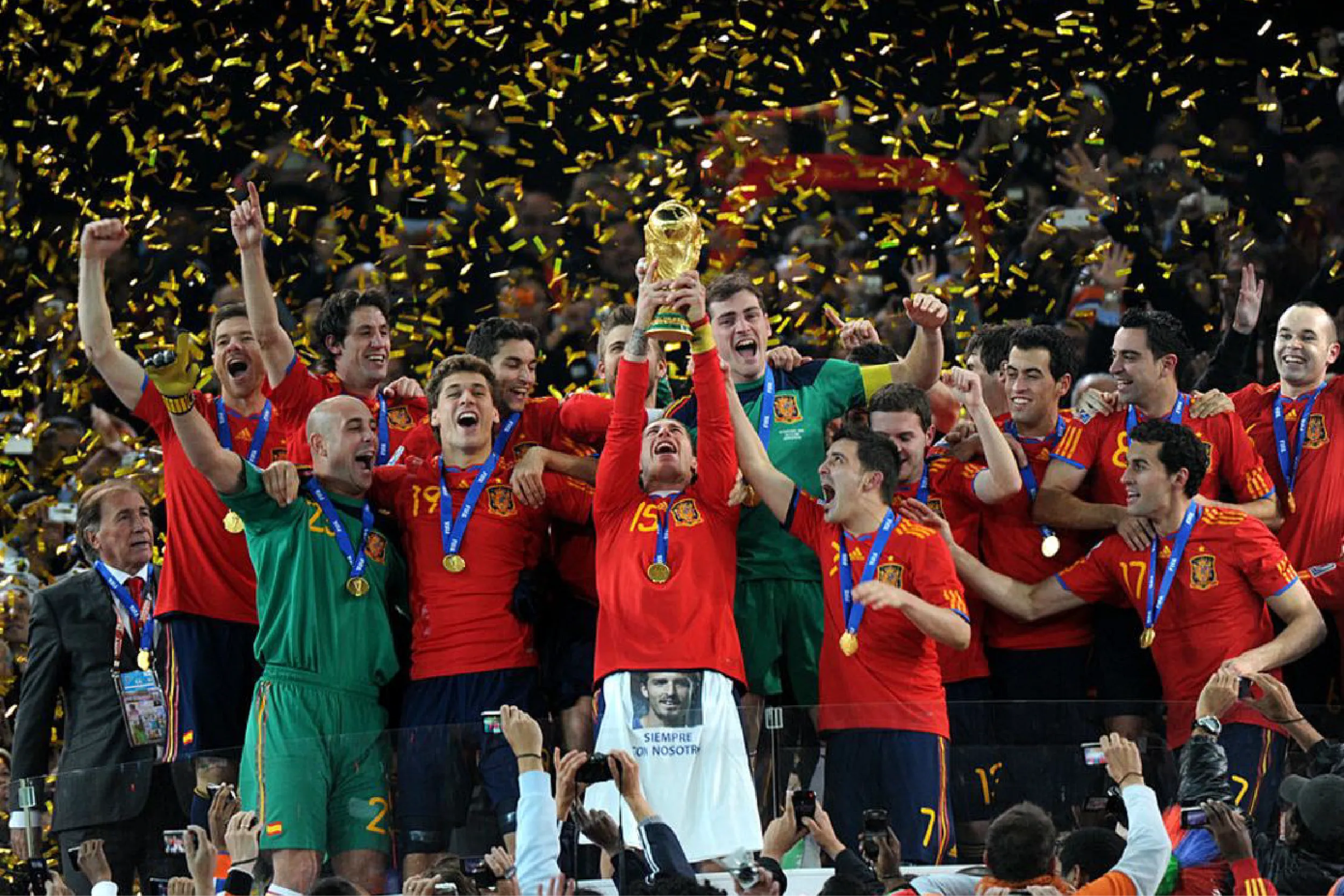 Spain national soccer team celebrating with trophy, wearing red uniforms and gold medals as gold confetti falls overhead