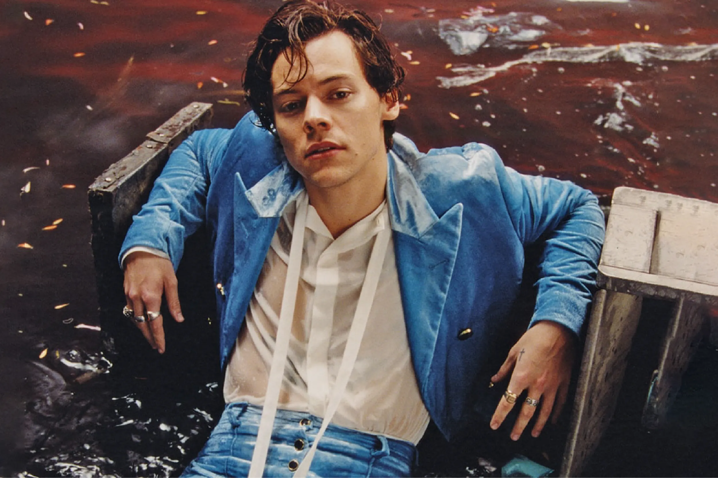 Man in a blue denim jacket with a white shirt and scarf sits in a small wooden boat on dark red water, looking toward the camera.
