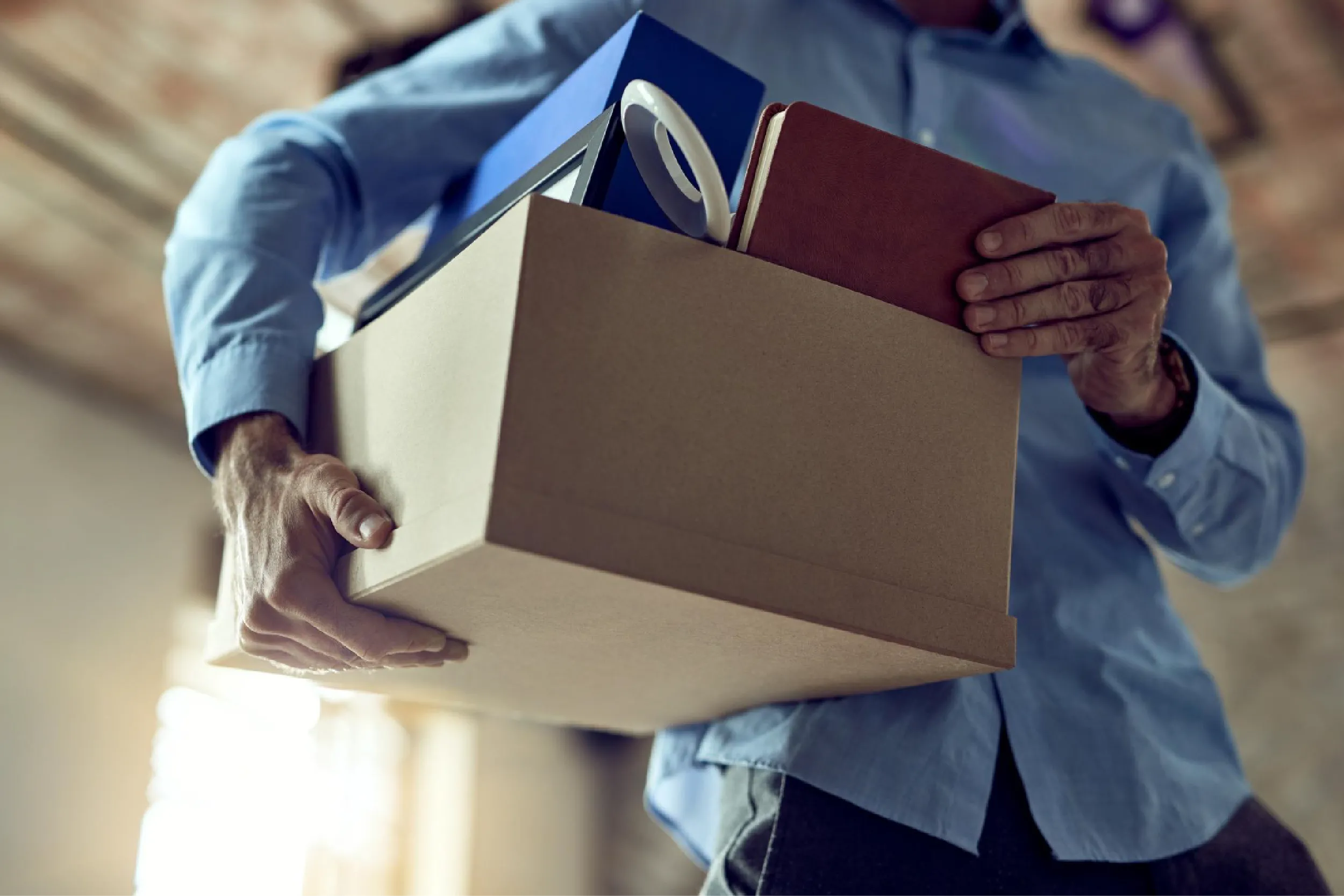 Person in a blue shirt carries a cardboard box with smaller boxes and packing tape inside a home setting.