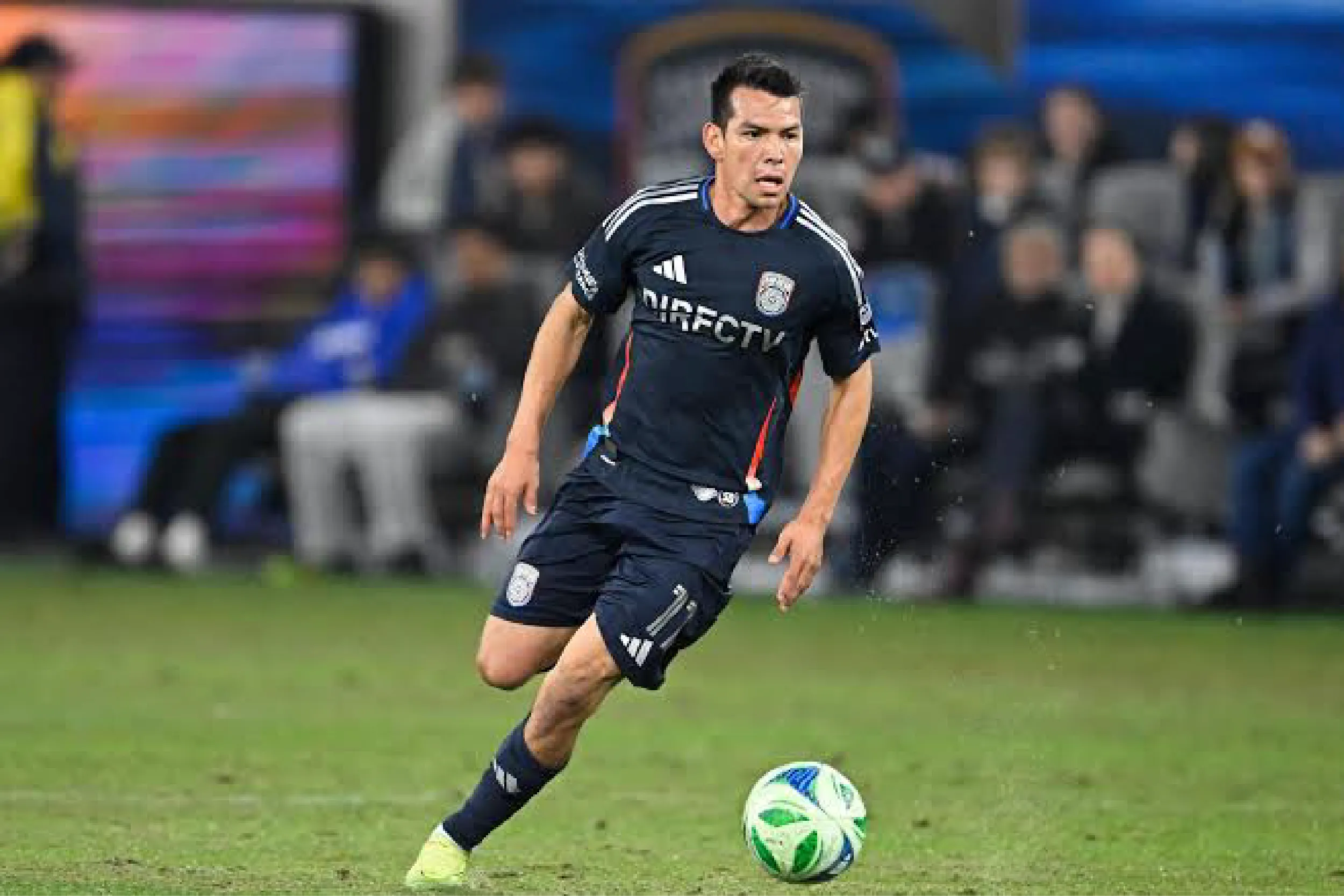 Soccer player in a dark navy jersey dribbling a green-and-white ball on a grass field during a match with blurred spectators in the background.