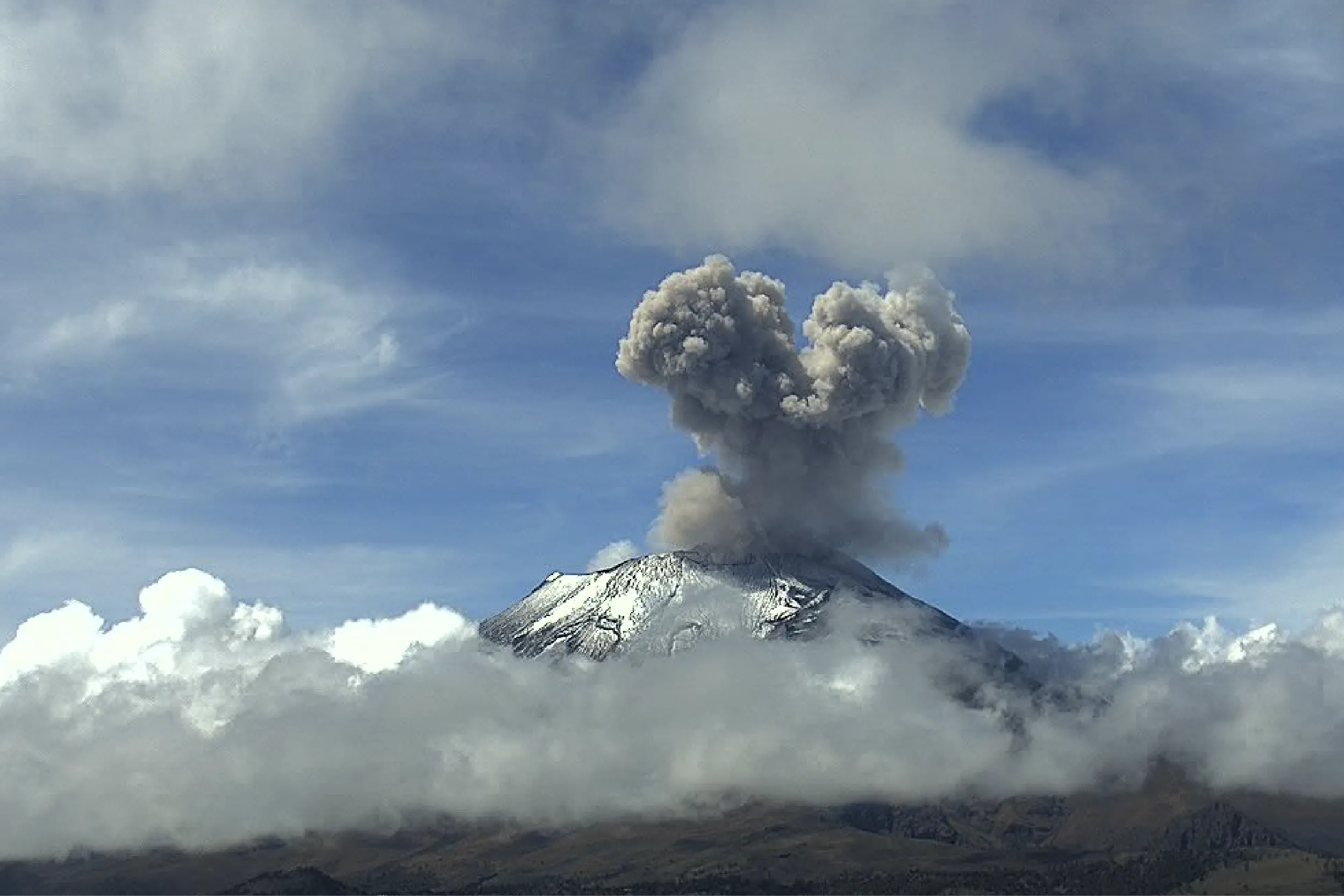 Snowy volcano erupts with a tall ash plume rising into the blue sky, surrounded by clouds