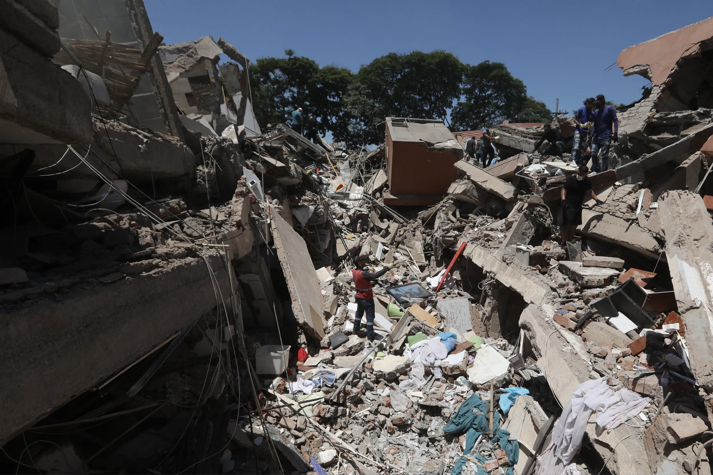 Rescue workers navigate a massive rubble field of collapsed buildings under a clear blue sky, searching for survivors amid debris.