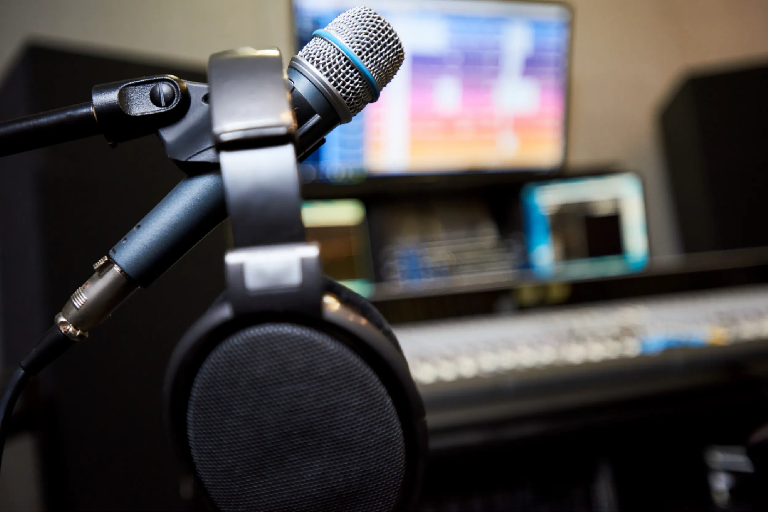 Dynamic microphone on an adjustable boom arm with over-ear headphones in the foreground and blurred computer screens in the background, suggesting a recording studio setup.